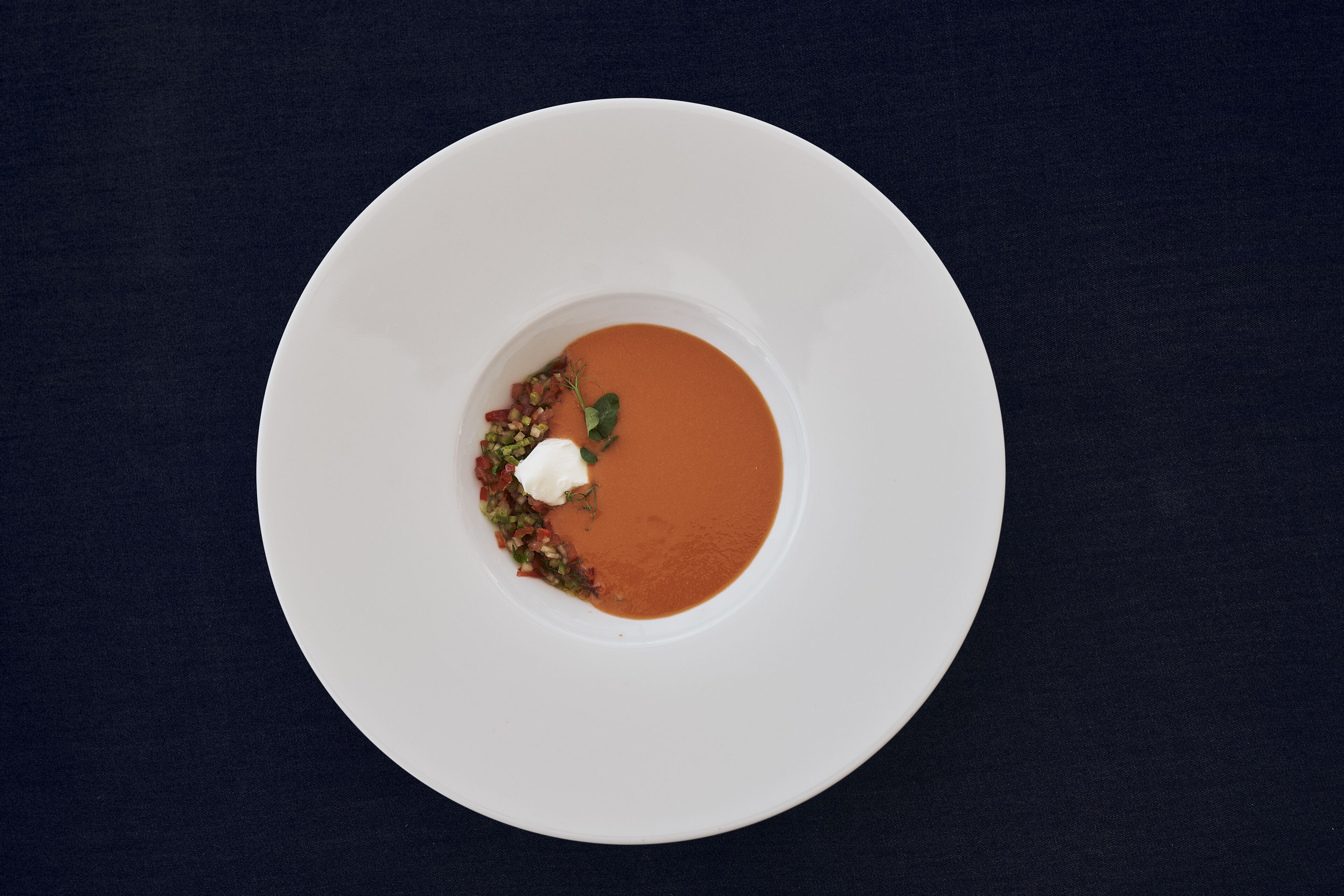 Une assiette blanche contenant un bol de soupe, agrémentée d’une cuillerée de crème et d’herbes hachées, posée sur une surface sombre.