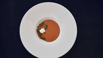 Une assiette blanche contenant un bol de soupe, agrémentée d’une cuillerée de crème et d’herbes hachées, posée sur une surface sombre.