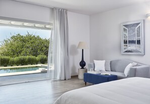 A hotel room with a large bed, a sofa, a small table, a lamp, and a painting. There is a window with a view of a pool and greenery outside. - Yria Boutique Hotel & Spa (Paros)