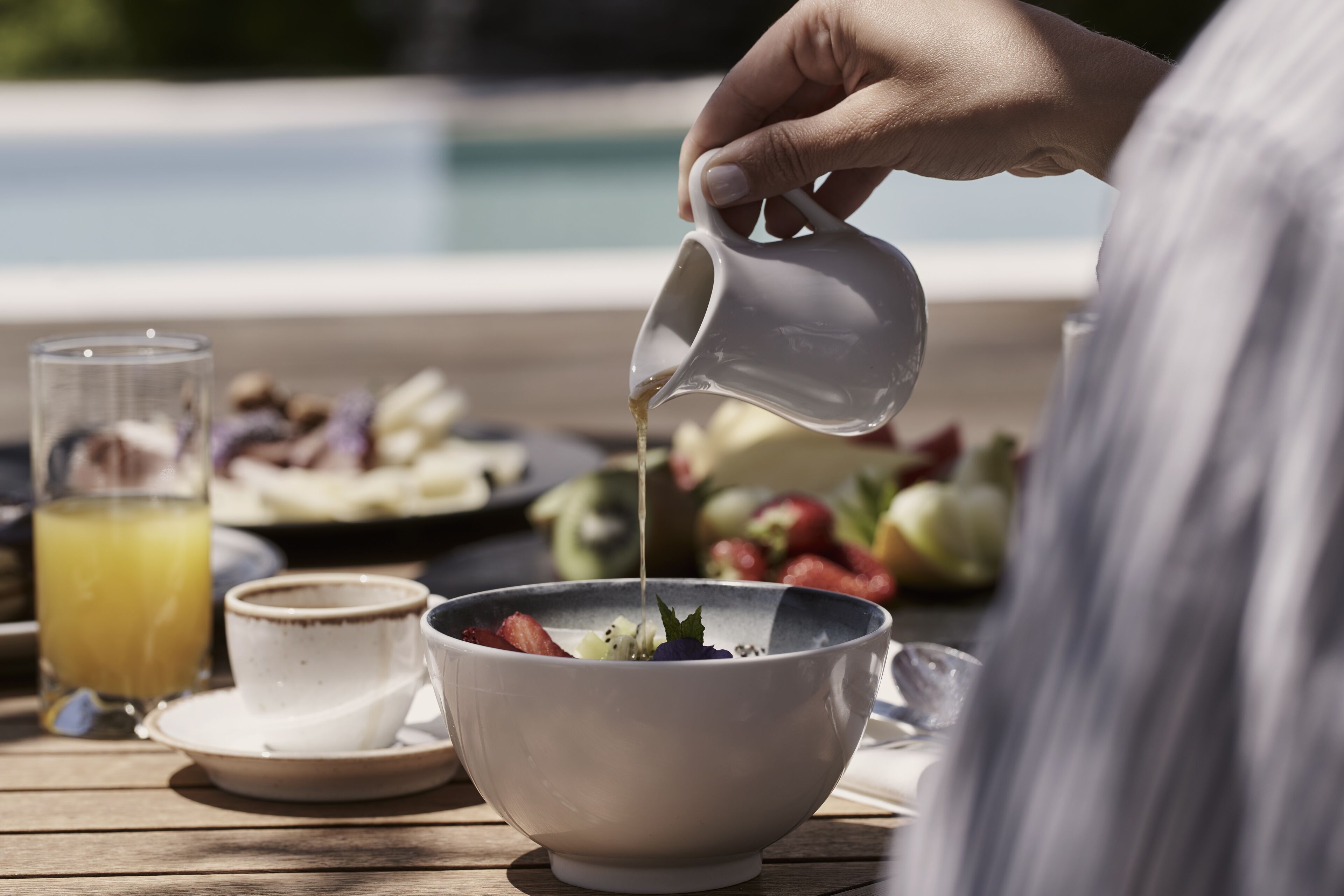 A person pouring a liquid into a bowl on a wooden table with a glass of orange juice and a plate of food in the background.