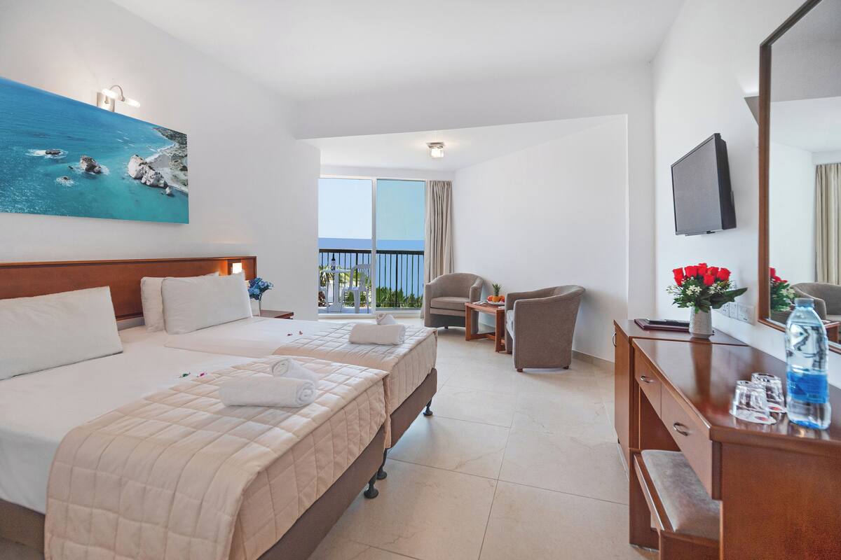 A hotel room with a bed, a desk, a chair, a TV, and a balcony with a view of the sea.