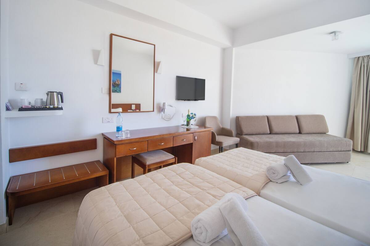 A hotel room with two beds, a sofa, a desk with a mirror, a television, and a small table with a kettle and cups.