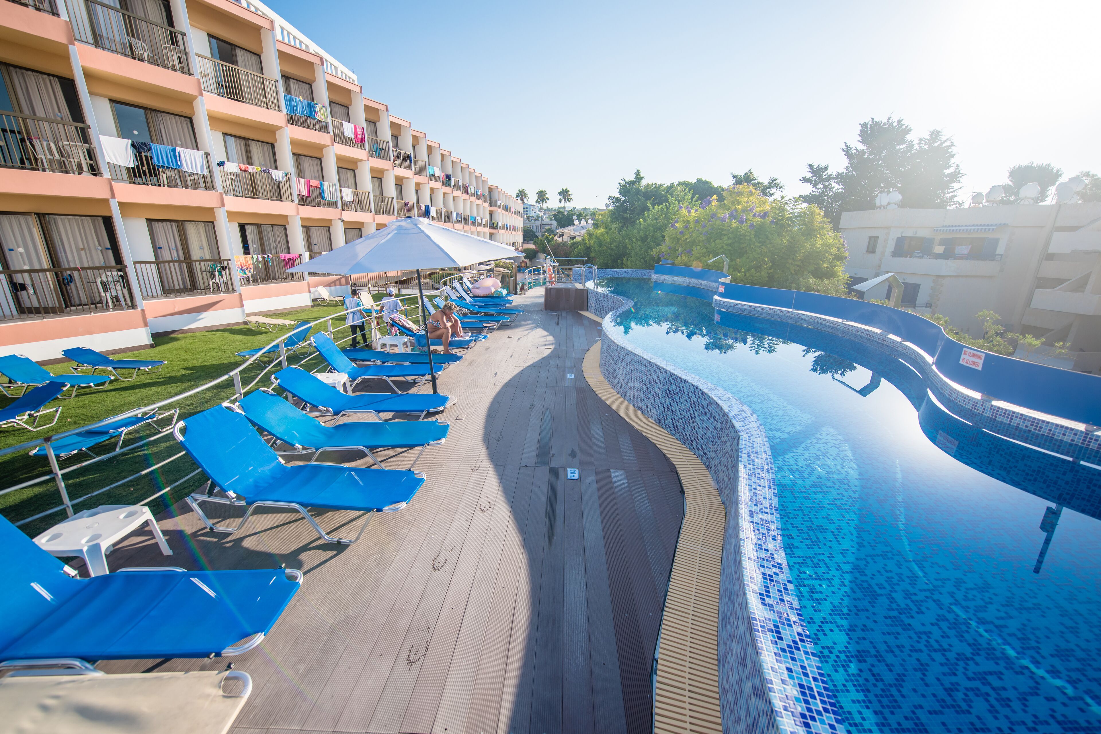 Un área junto a la piscina con sillas reclinables azules y sombrillas, una terraza de madera y un edificio con balcones.