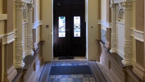 Un amplio pasillo con columnas blancas ornamentadas, una puerta negra con vidrieras y una alfombra estampada.