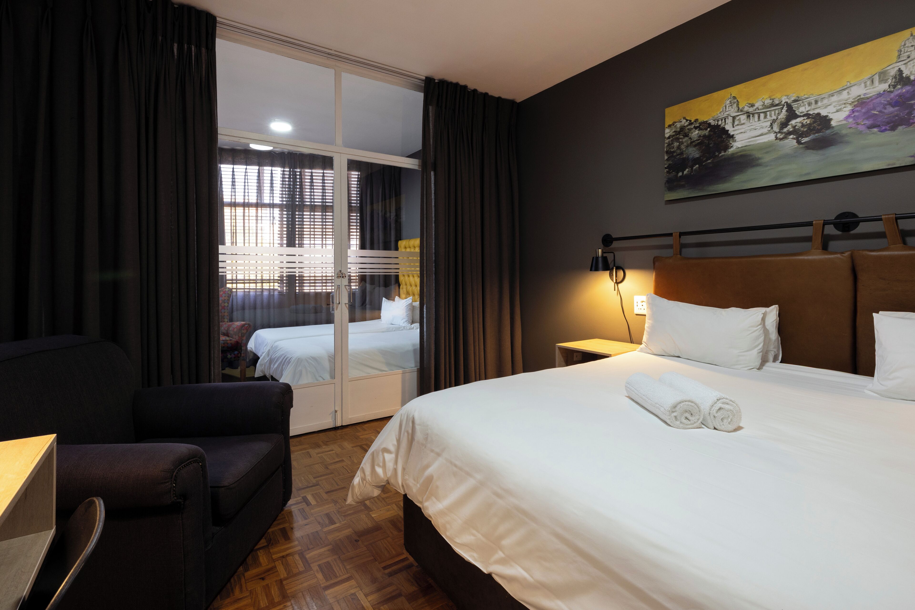 A hotel room with a large bed, a desk, a chair, and a view of another room through a glass door.