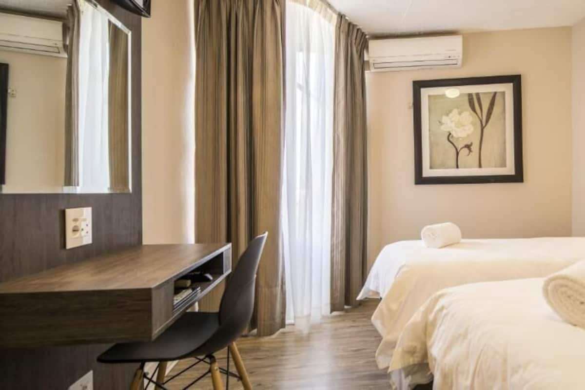 Een hotelkamer met twee bedden, een bureau, een stoel, een groot raam met gordijnen, en een ingelijst schilderij aan de muur.