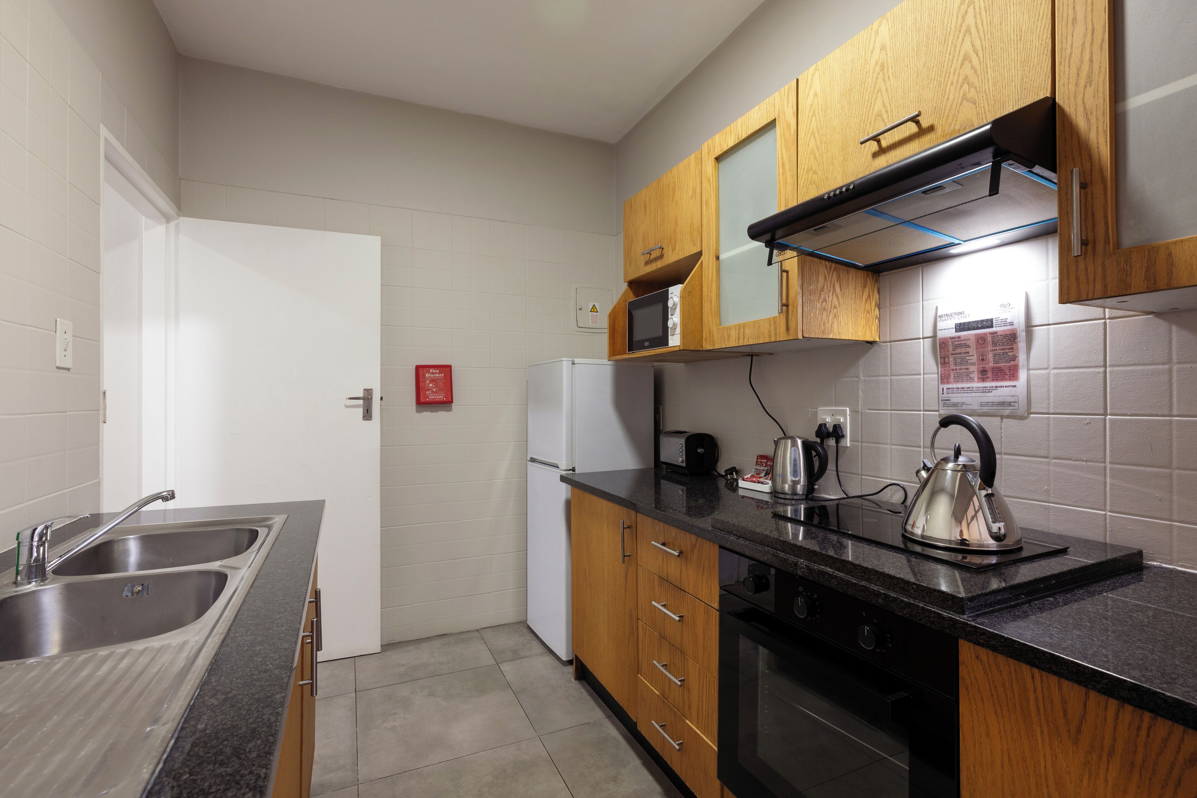 A compact kitchen with a stainless steel sink, wooden cabinets, a microwave, and a kettle.