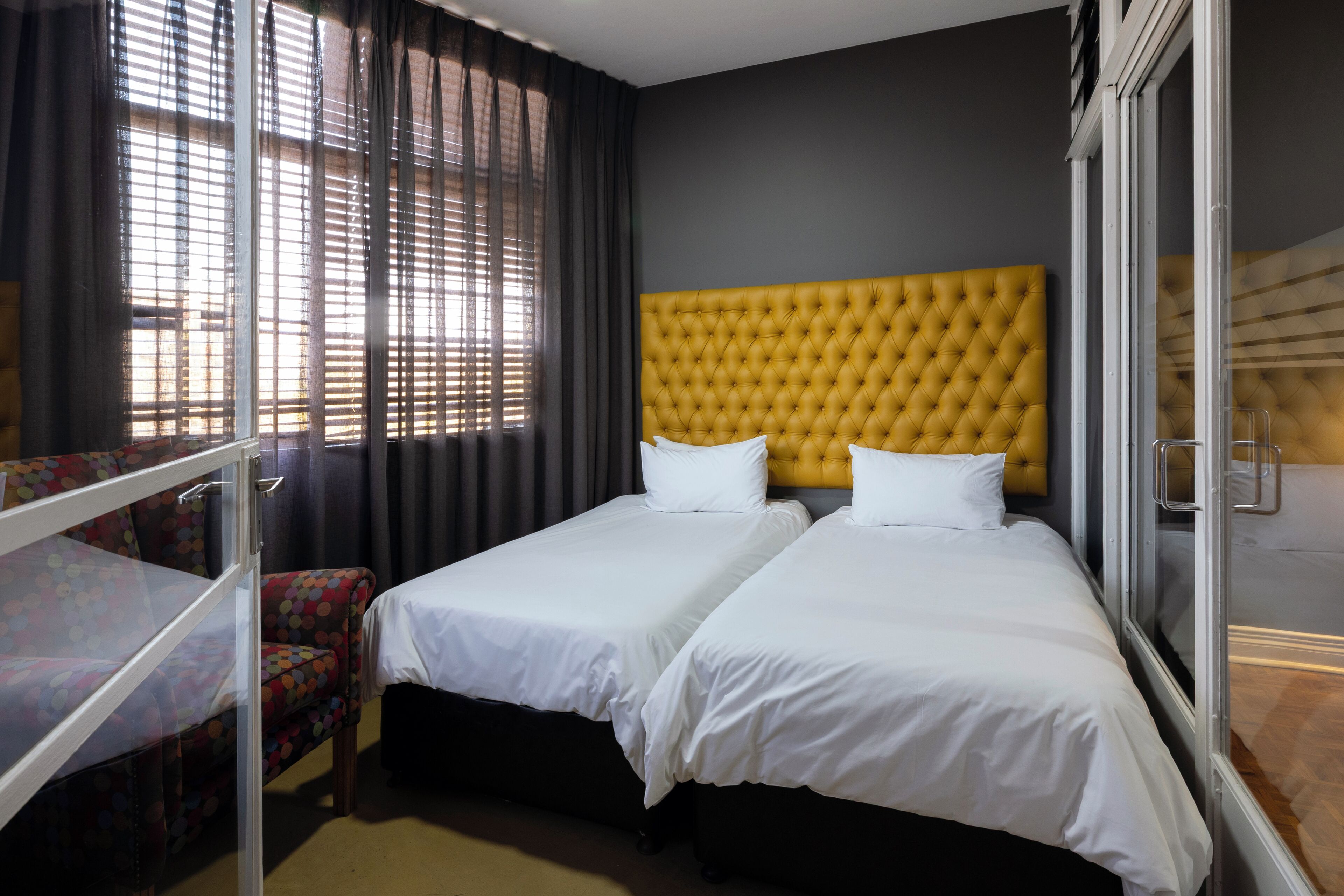 Ein Hotelzimmer mit zwei Betten, einem Fenster mit Jalousien, einem Sessel und einem Bett mit Kopfteil.