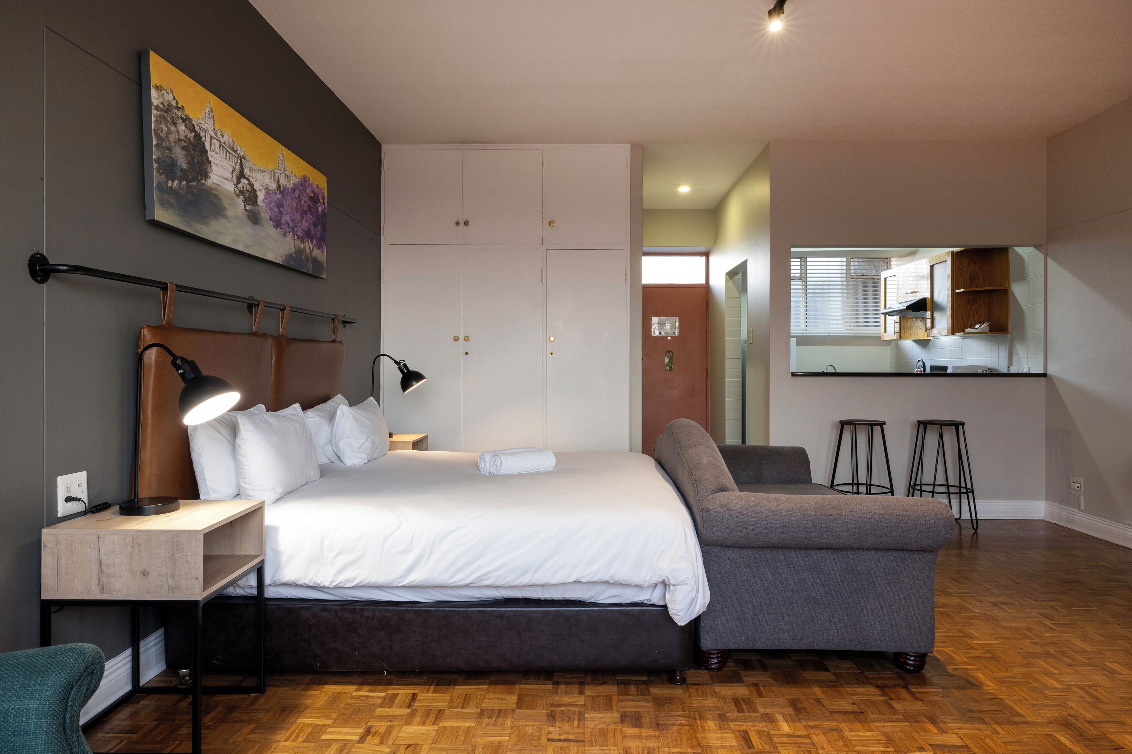 Ein modernes Schlafzimmer mit einem großen Bett, Nachttischen, einem Schreibtisch, einem Sessel, einer Couch und einer kleinen Küchenzeile mit Sitzhockern.