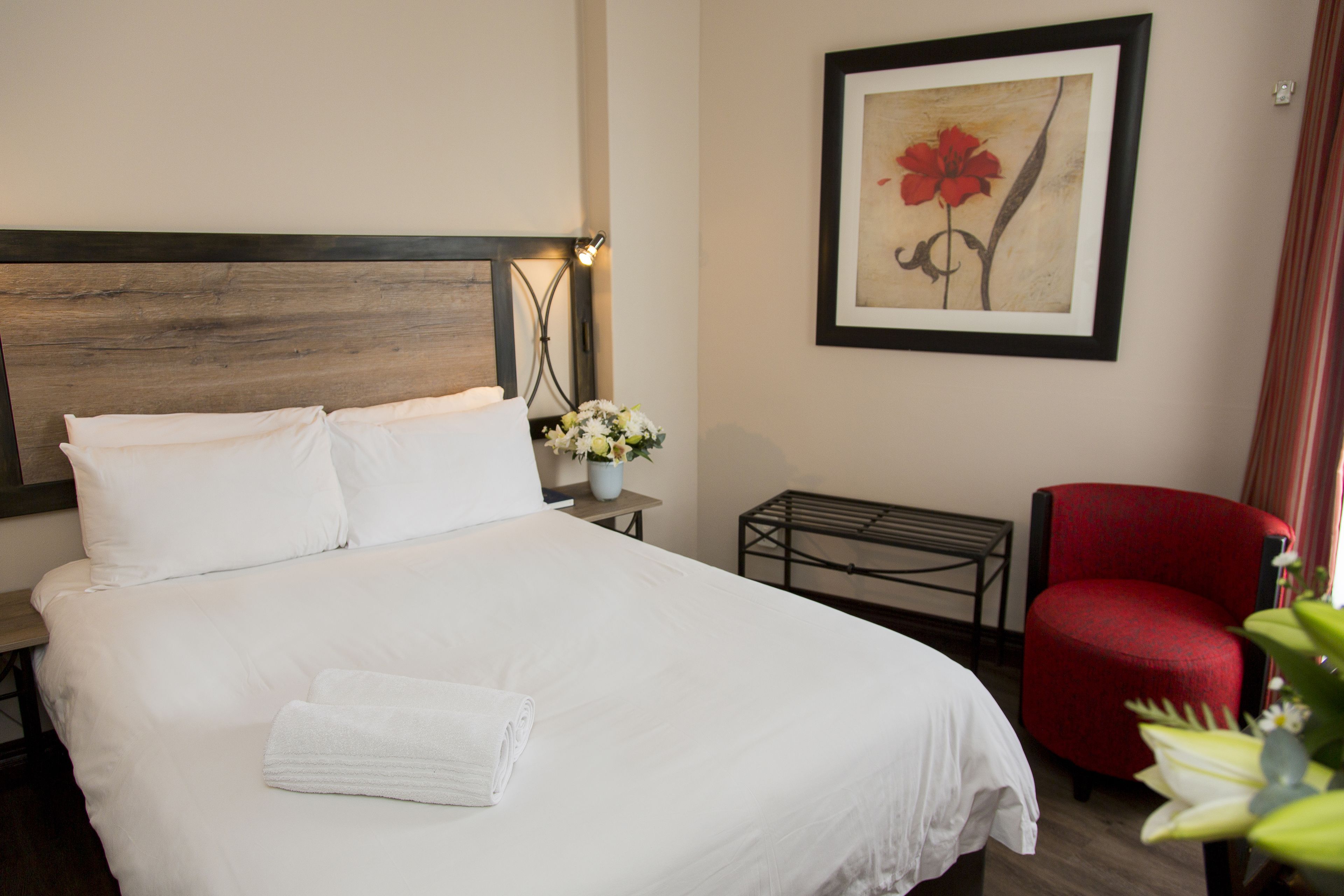 Una cama bien hecha con sábanas blancas y una toalla doblada, una mesita con un florero, una silla roja y una obra de arte enmarcada en la pared.