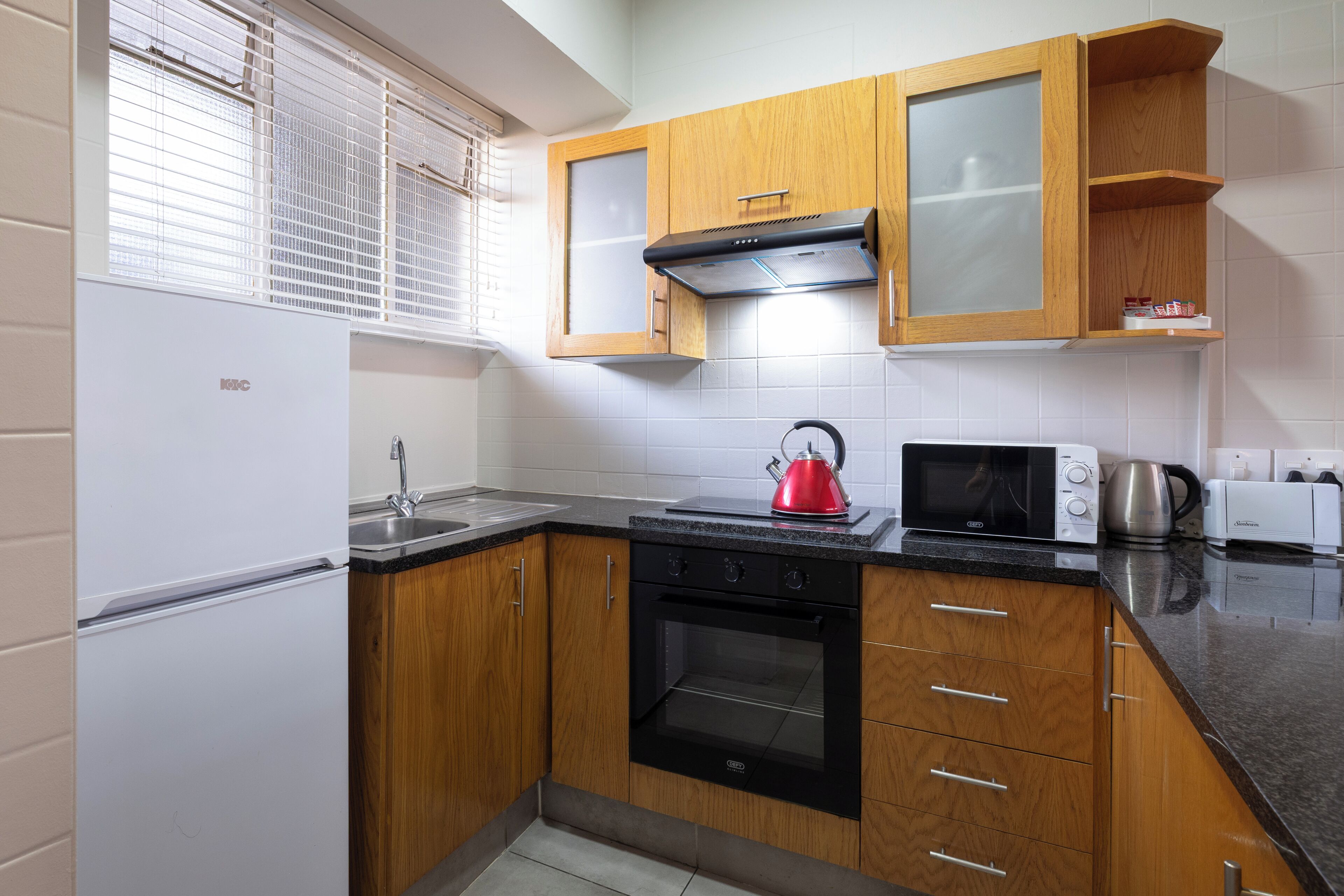 A compact kitchen with wooden cabinets, a stainless steel sink, a black oven, a microwave, and a kettle.