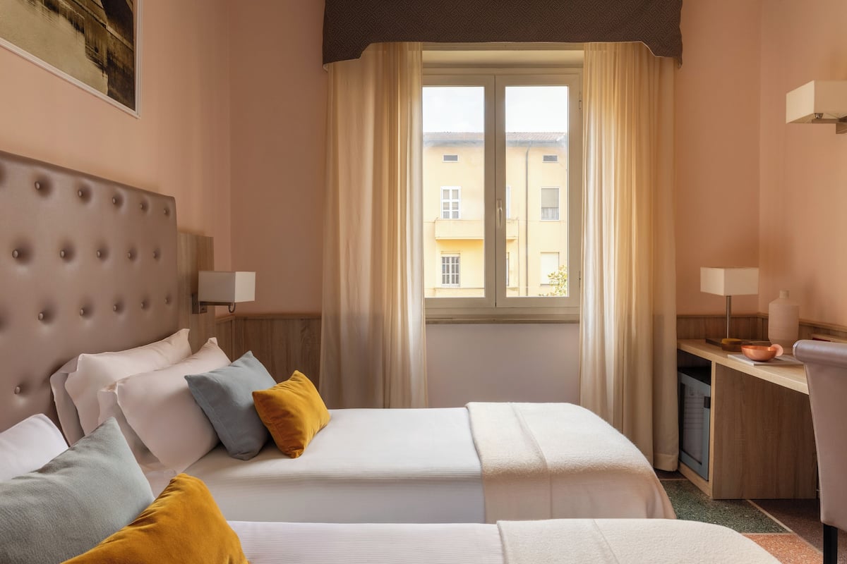 Ein Hotelzimmer mit einem Bett, einem Schreibtisch, einem Stuhl, einem Fenster mit Vorhängen und Blick auf Gebäude.