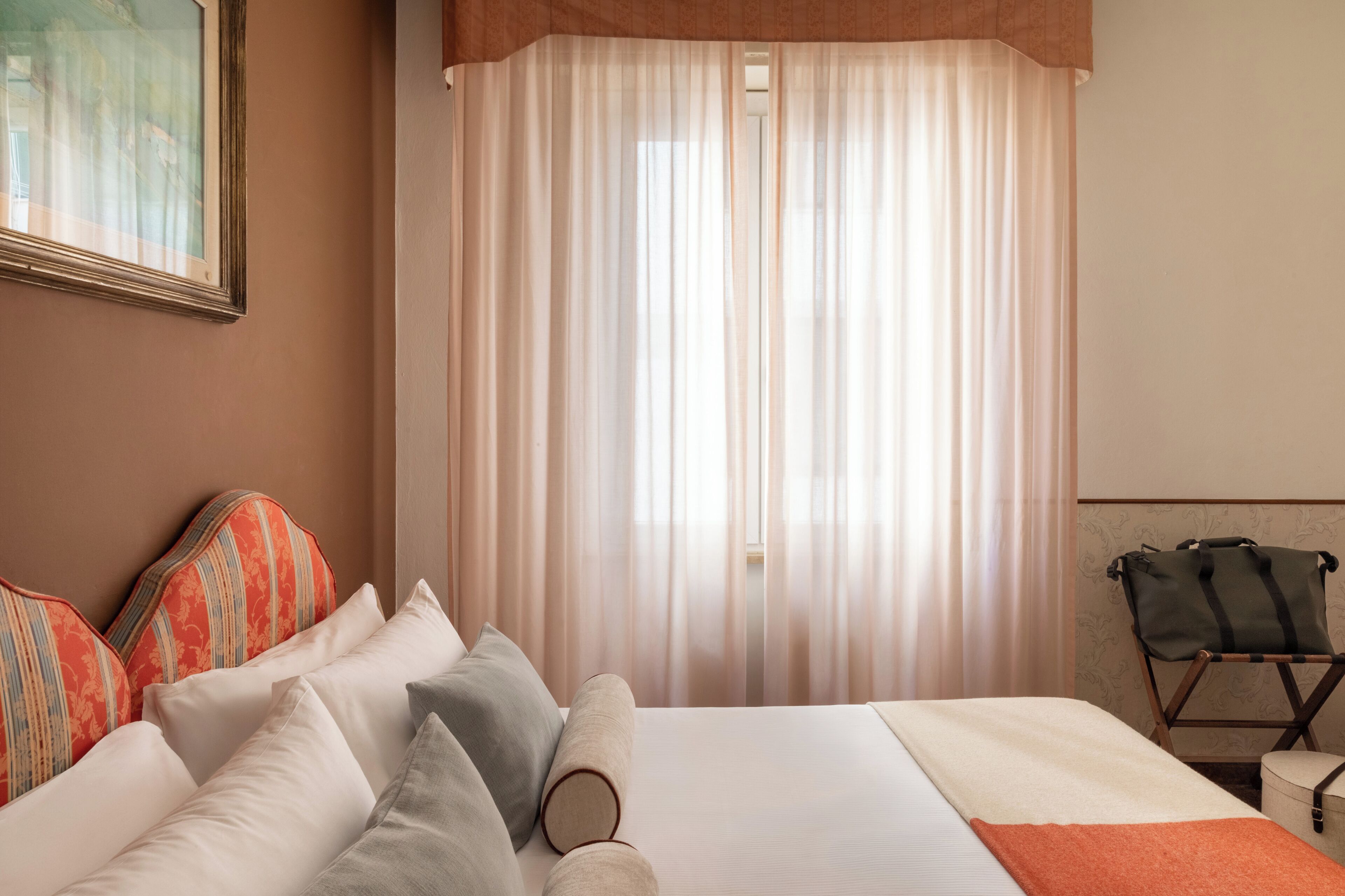 Una camera d'albergo con un letto, cuscini, una valigia e una finestra con tende leggere.