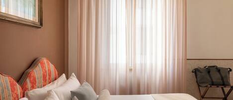 A hotel room with a bed, pillows, a suitcase, and a window with sheer curtains.