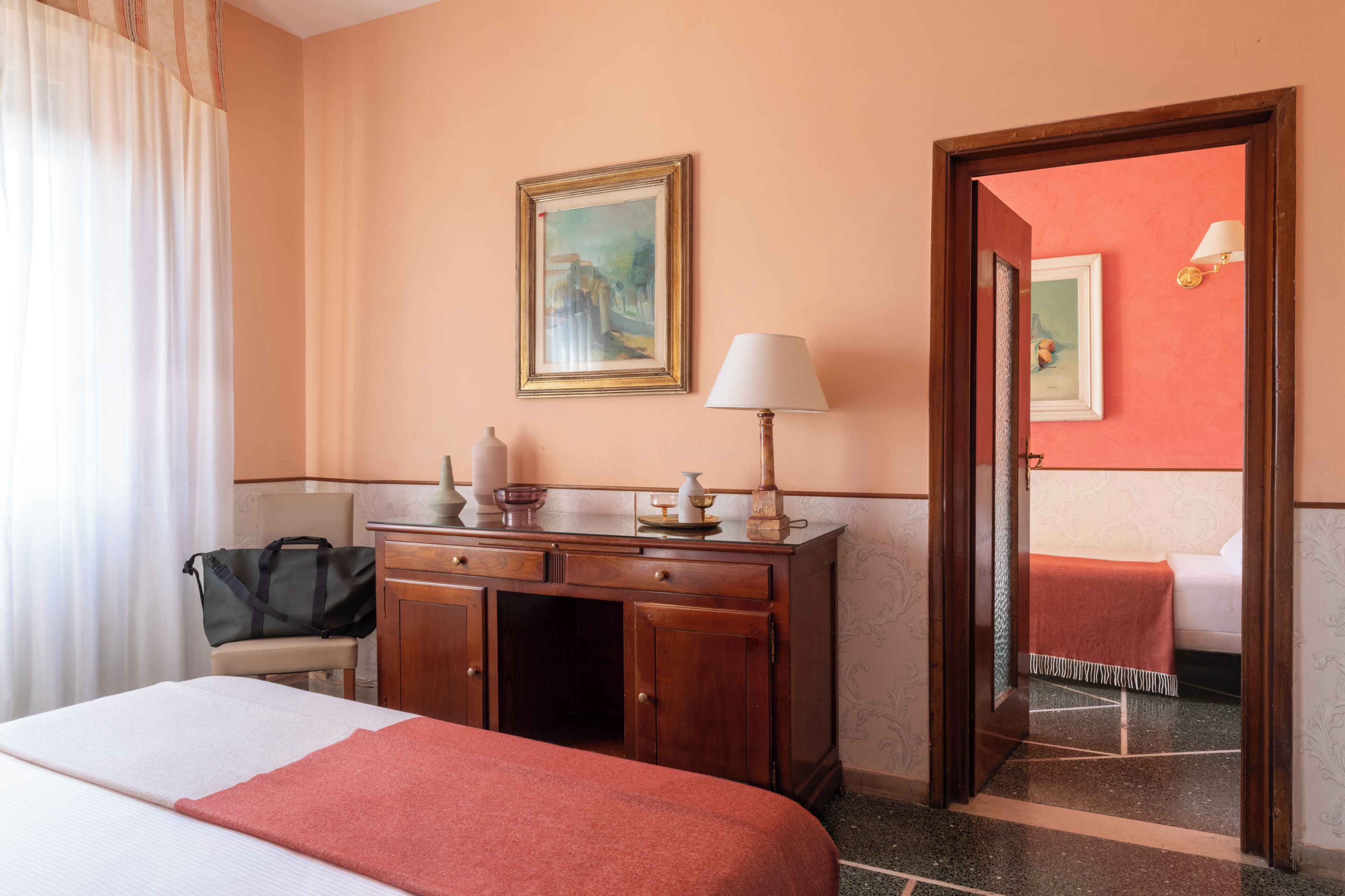 A hotel room with a bed, a wooden dresser, a desk, a lamp, a painting on the wall, and a mirror.
