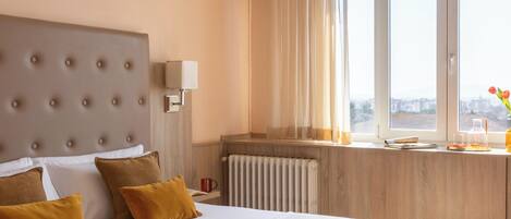 Habitación de hotel con una cama grande, cabecero, ventana con cortinas, radiador y un cuadro en la pared.