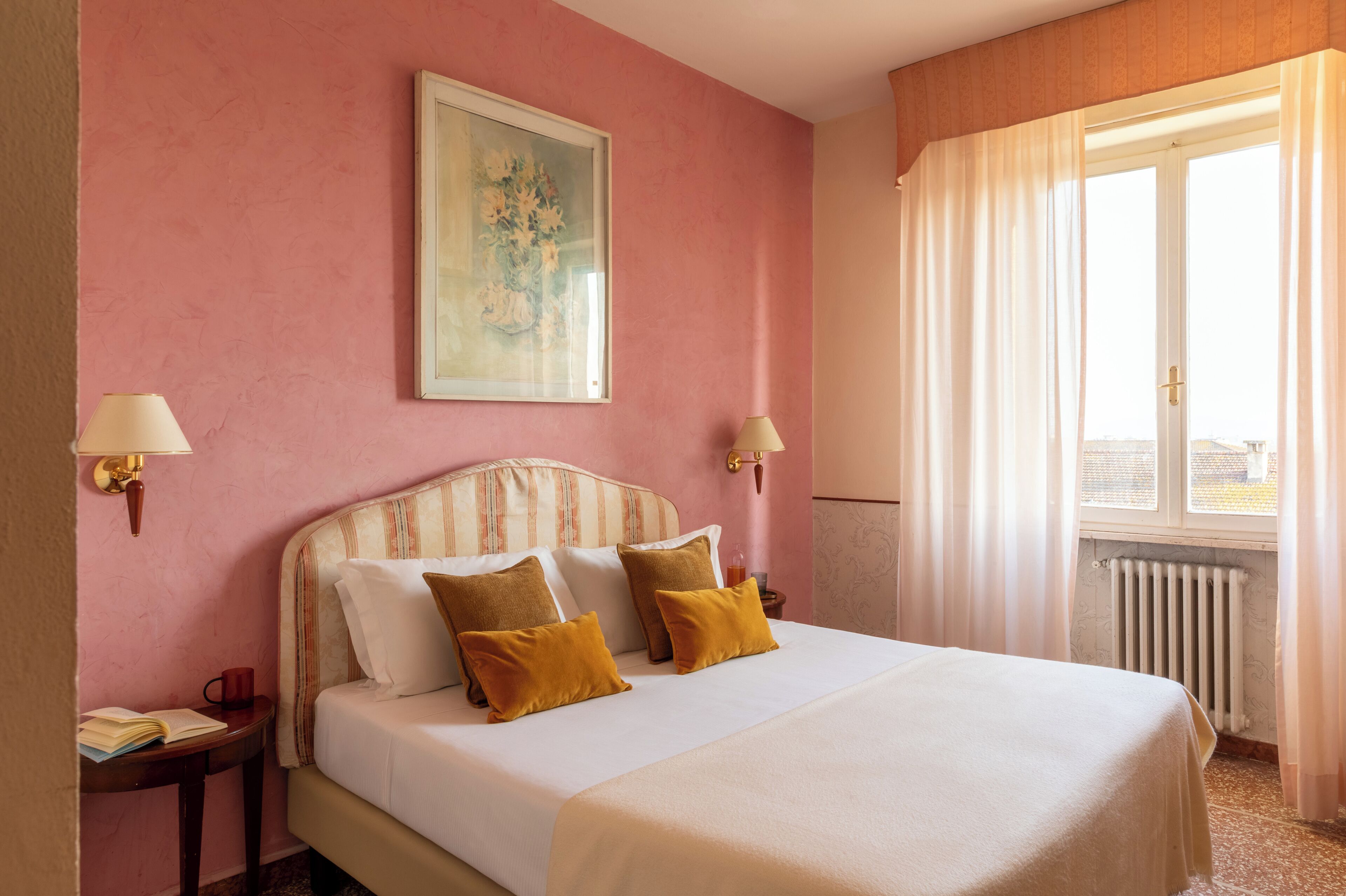 A hotel room with a bed, two bedside tables, a nightstand, a window with curtains, and a painting on the wall.