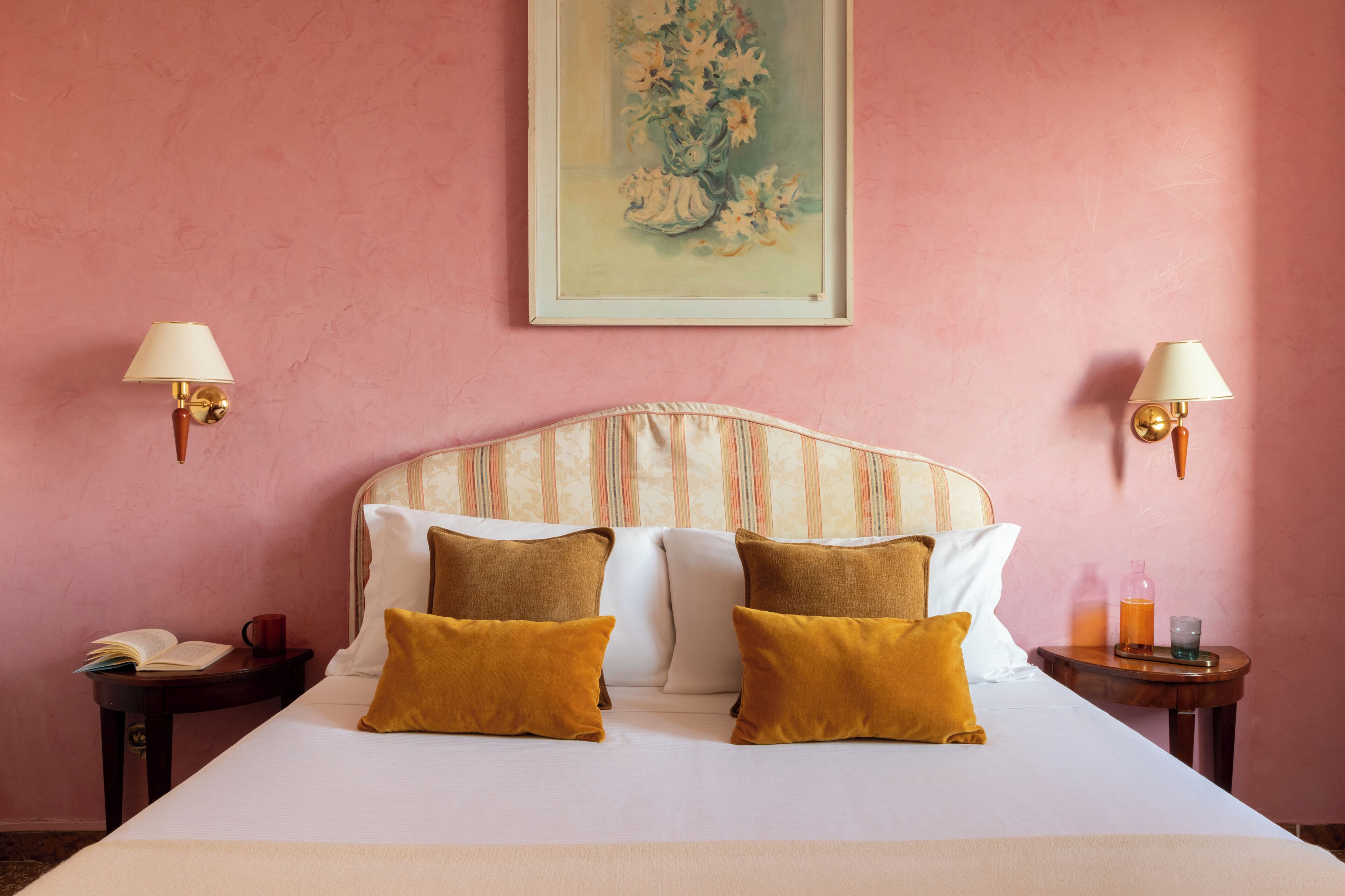 Una habitación de hotel con una cama, dos lámparas de noche, una mesita de noche con un libro y una taza, y un cuadro en la pared.