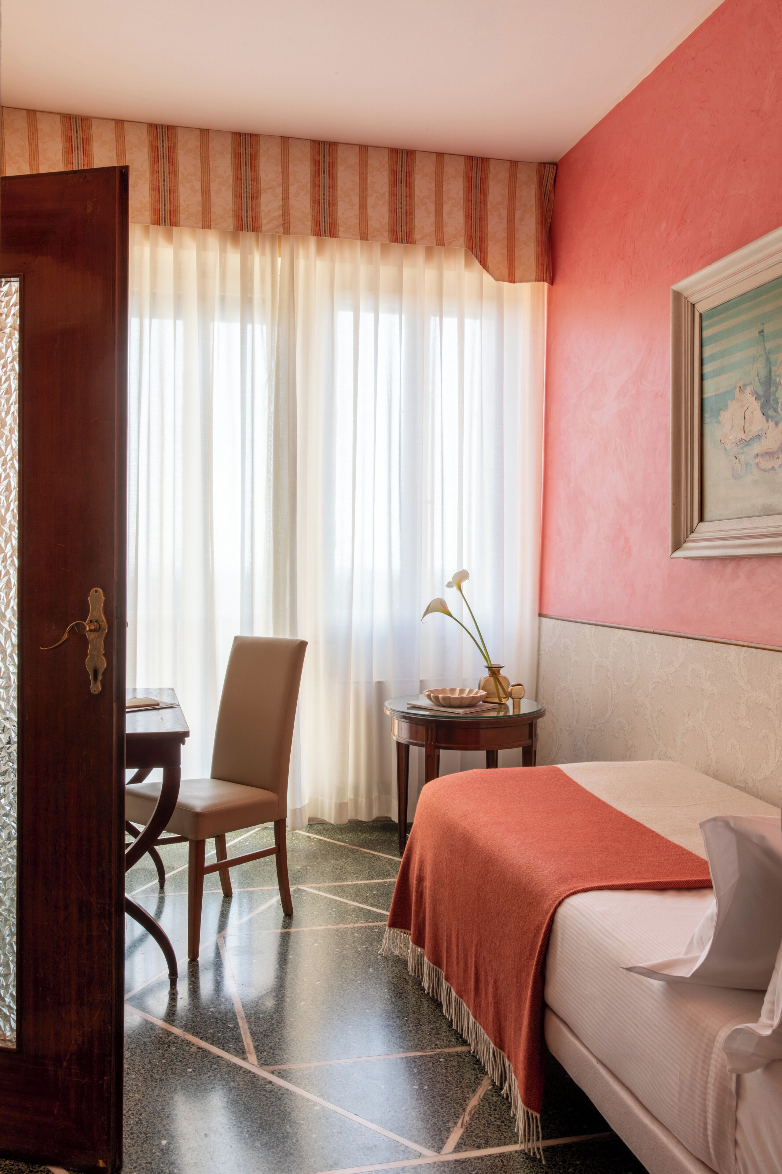 A hotel room with a bed, a chair, a small table with a vase, and a painting on the wall.