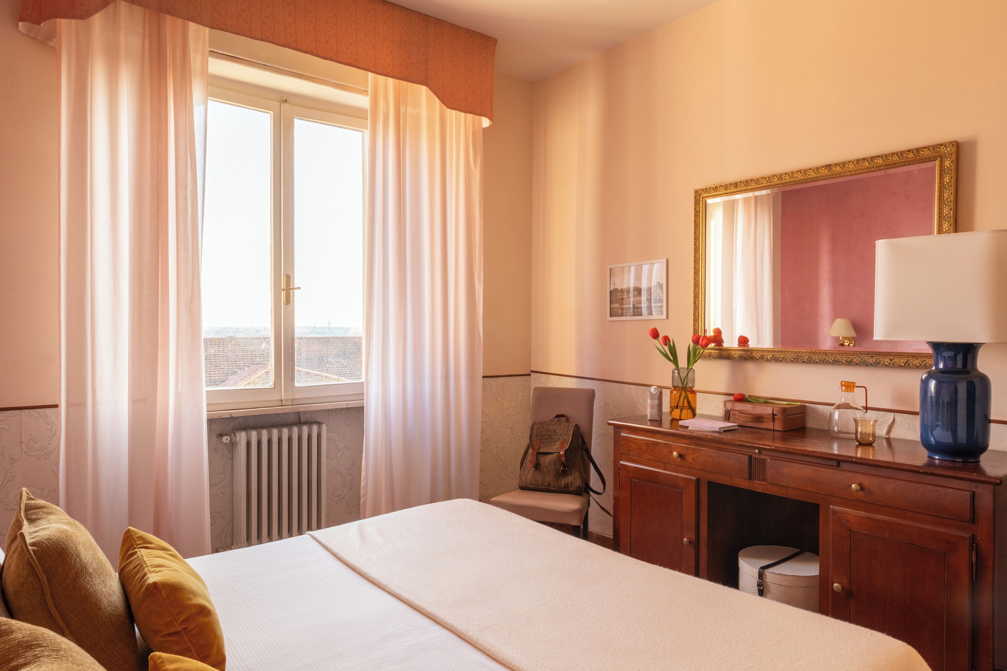 A hotel room with a bed, a desk, a chair, a radiator, a window with curtains, a mirror, and a vase with flowers.