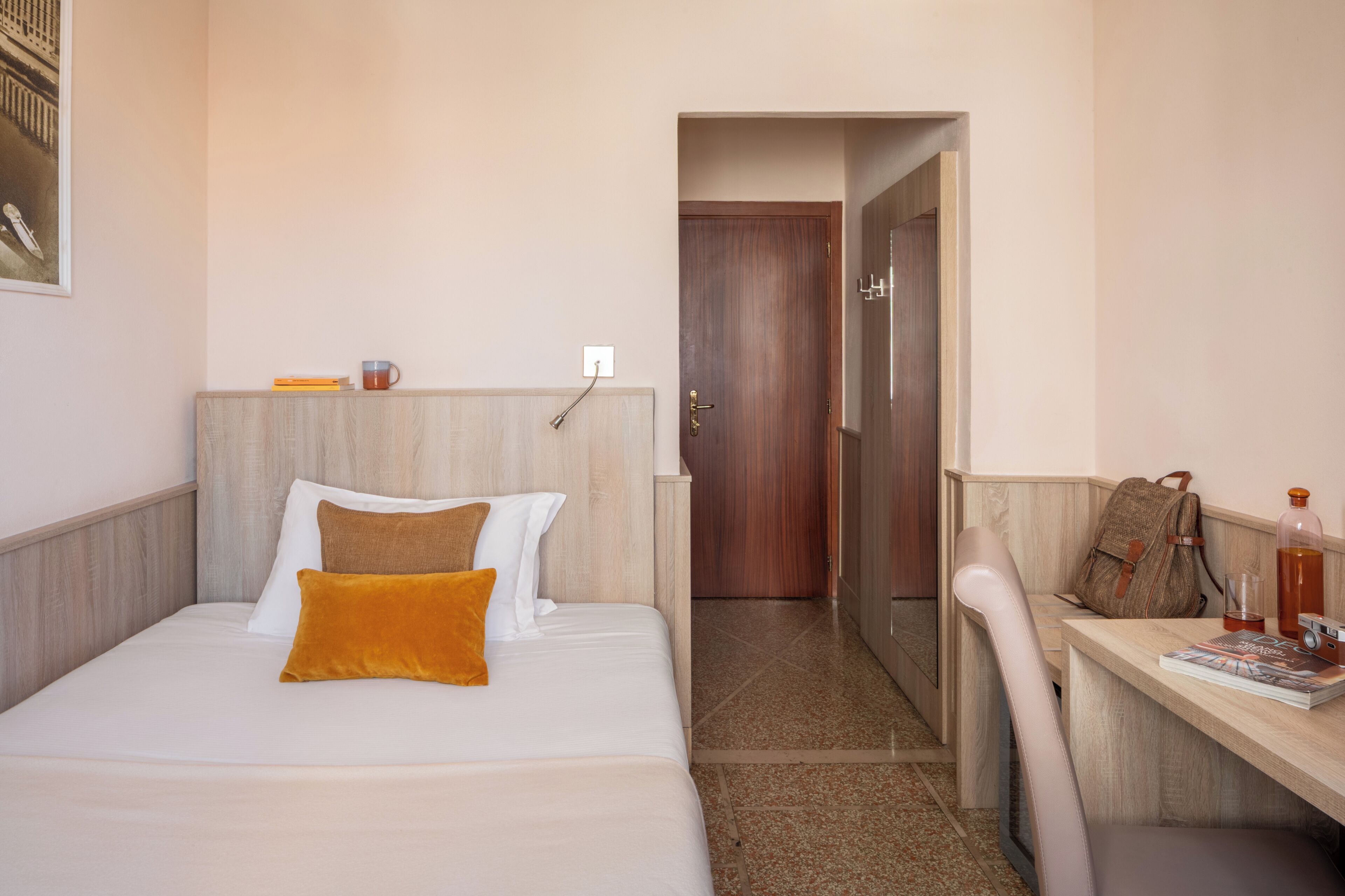 A hotel room with a bed, a desk, a chair, a door, and a wall-mounted shelf.