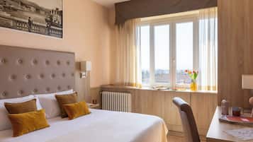 A hotel room with a large bed, a desk, a chair, a window with curtains, and a radiator.