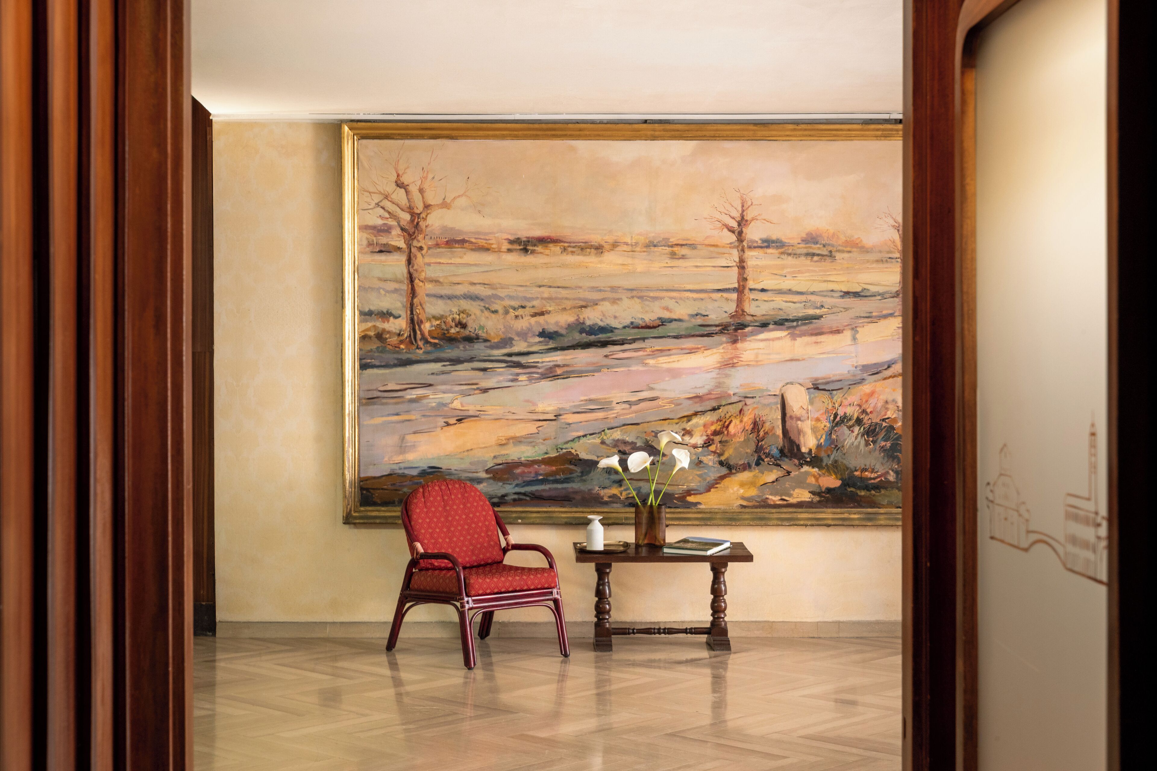 Une pièce décorée d’un grand tableau représentant un paysage, d’un fauteuil rouge recouvert de tissu, d’une petite table ornée d’un vase de fleurs et d’un sol carrelé.