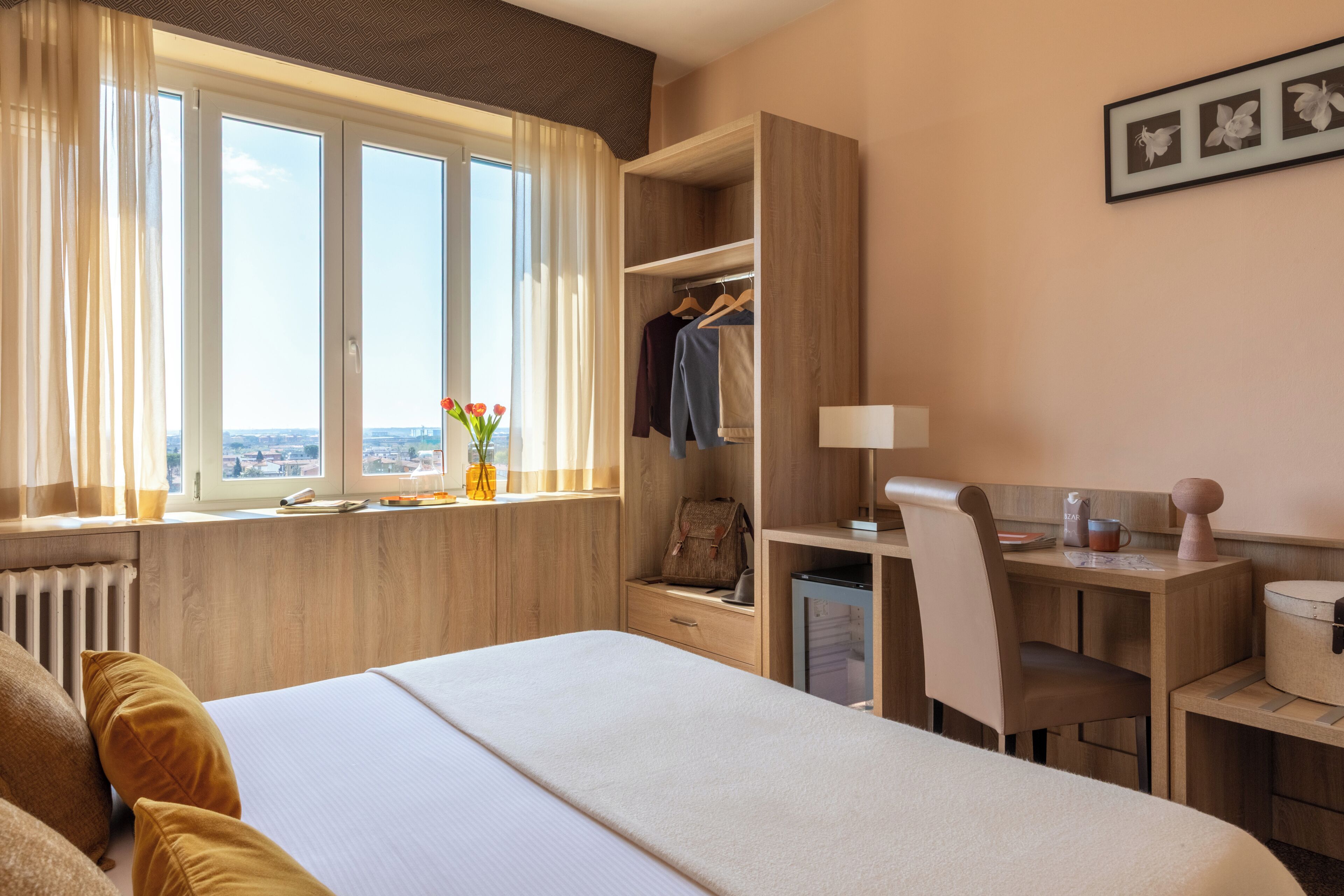 A hotel room with a bed, a desk, a chair, a radiator, a window with curtains, a view of the city, and a framed picture on the wall.