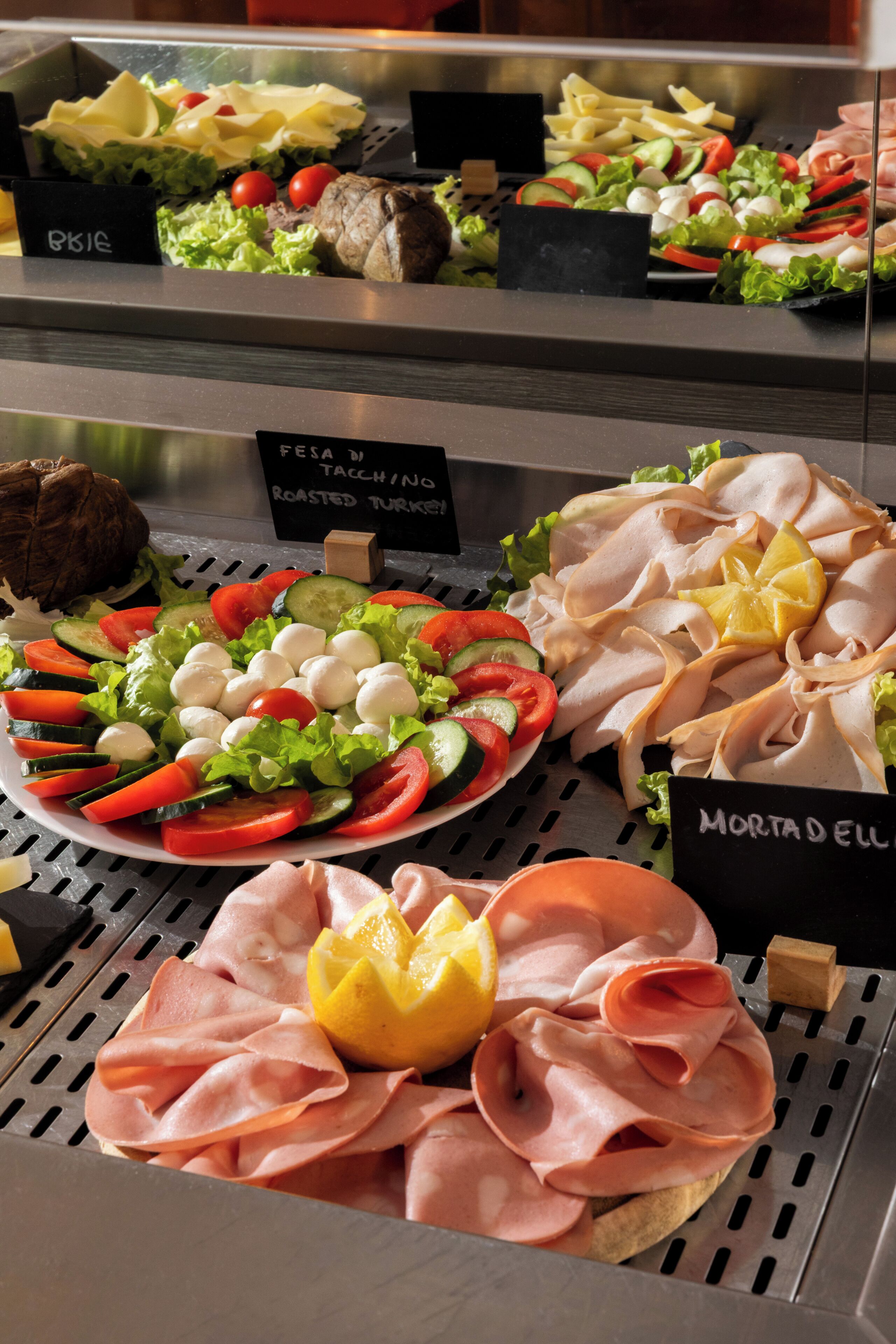 Diversos alimentos expuestos en una vitrina de charcutería, con etiquetas que identifican distintos tipos de carnes y ensaladas.