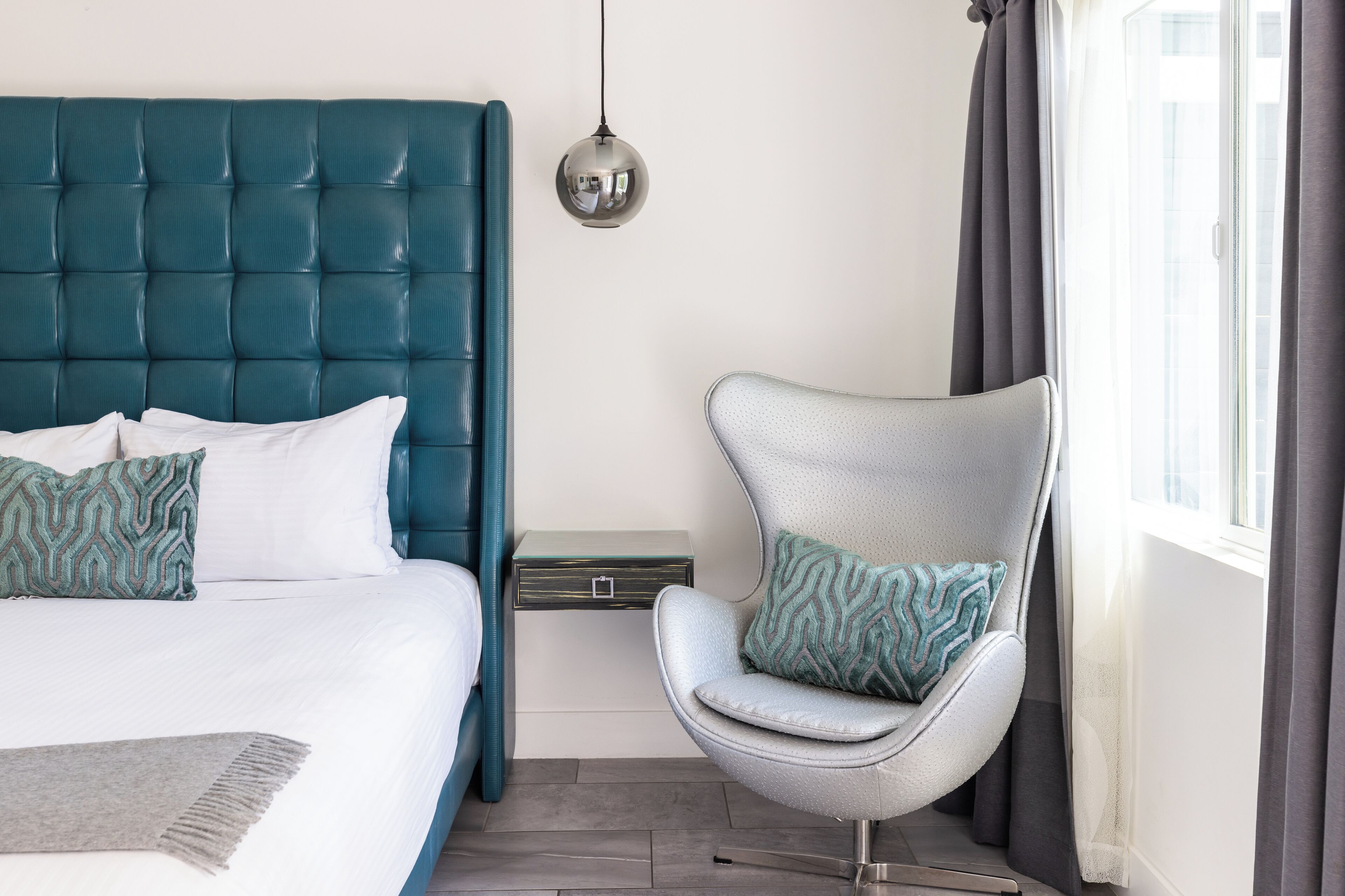 A modern bedroom with a teal upholstered headboard, a grey upholstered chair with patterned cushions, a small bedside table, and a hanging pendant light.