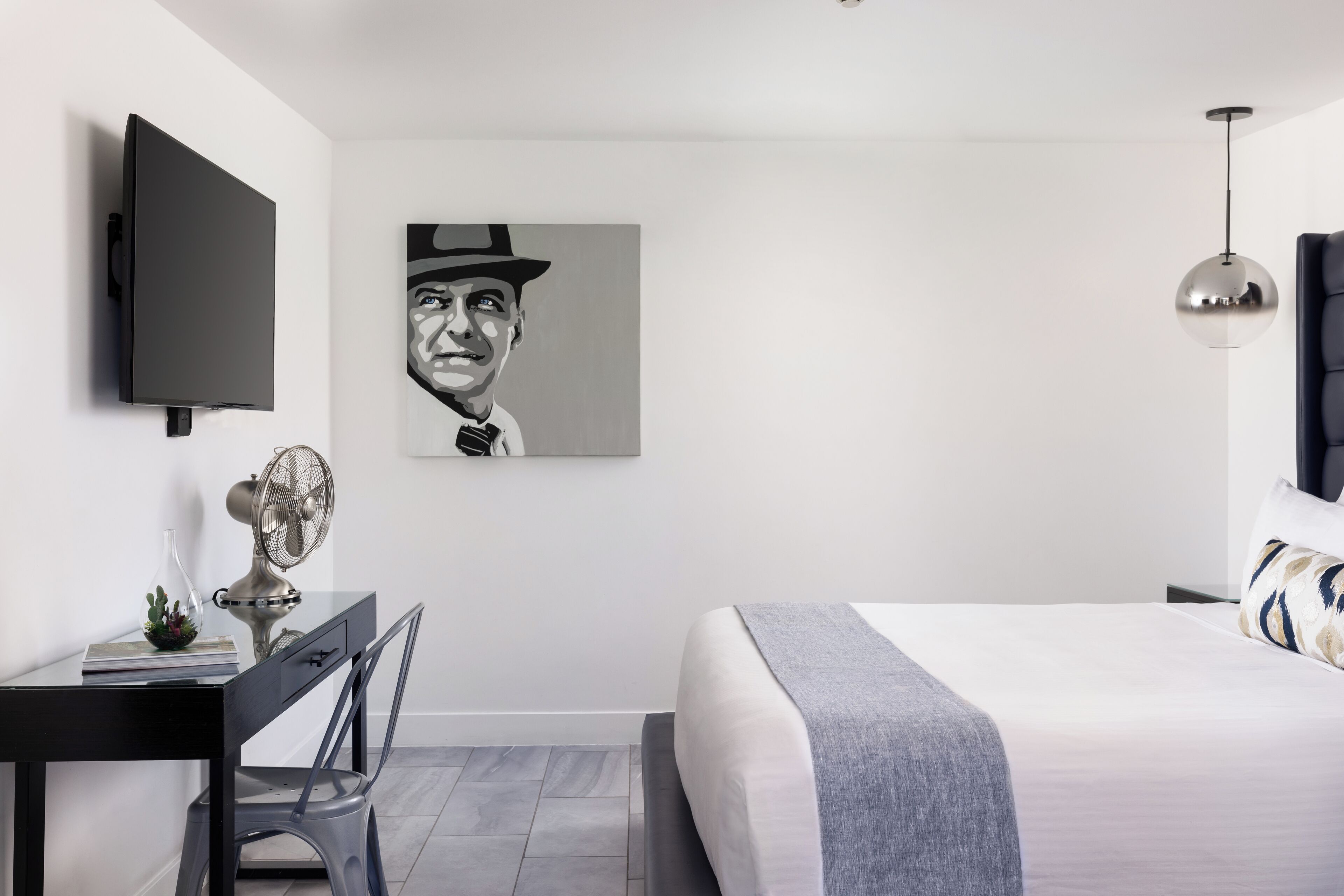 A modern bedroom with a bed, a desk with a chair, a television, and a wall-mounted artwork.