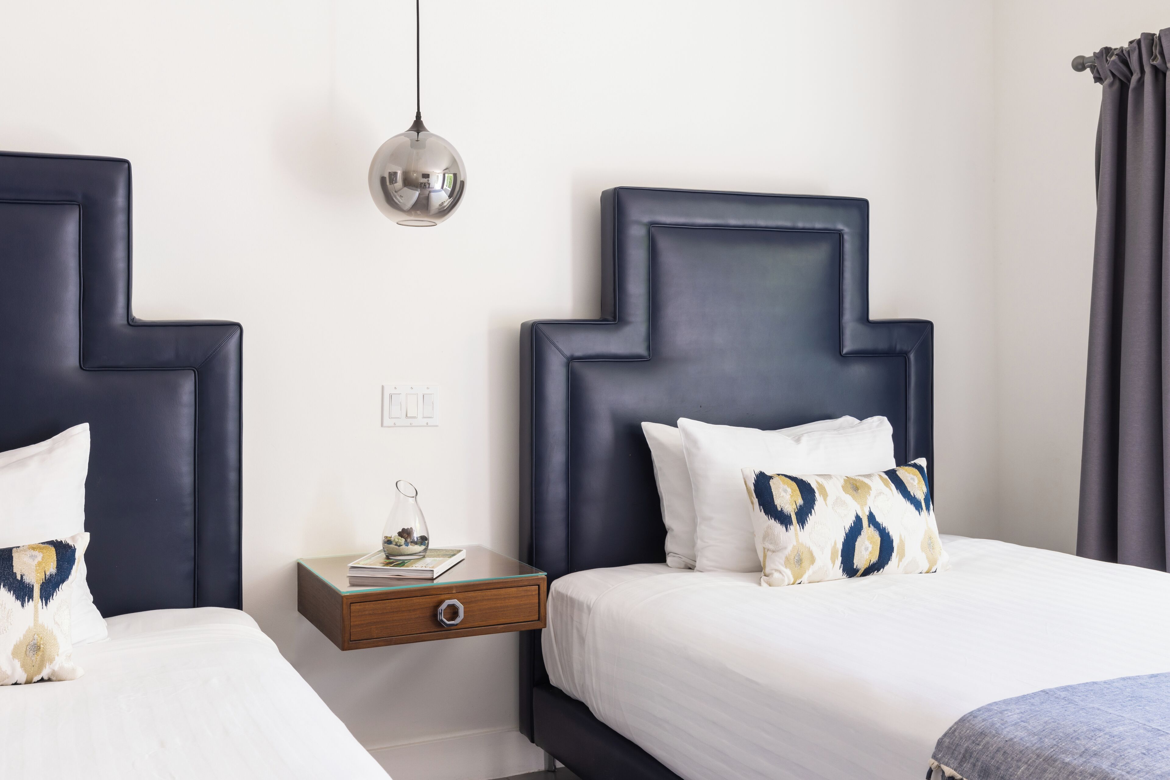 A modern hotel room with two beds, a nightstand, and a hanging pendant light.