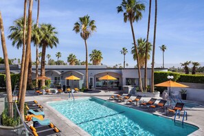 A swimming pool with lounge chairs and umbrellas, surrounded by palm trees and a building with arched windows. - The Palm Springs Hotel (Palm Springs)