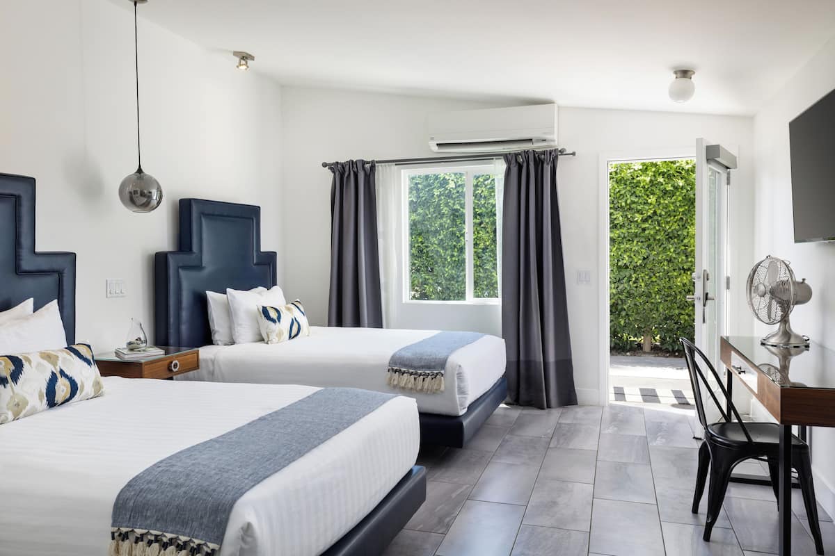 A hotel room with two beds, a desk, a chair, a fan, and a view of greenery outside.
