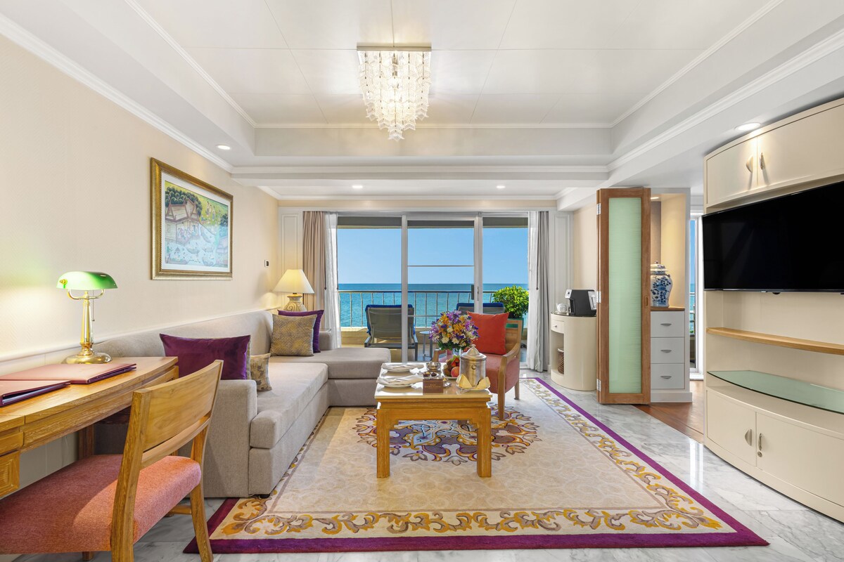 Un salon bien éclairé comprenant un coin repas, un canapé, un téléviseur et une vue sur la mer grâce à de grandes fenêtres.