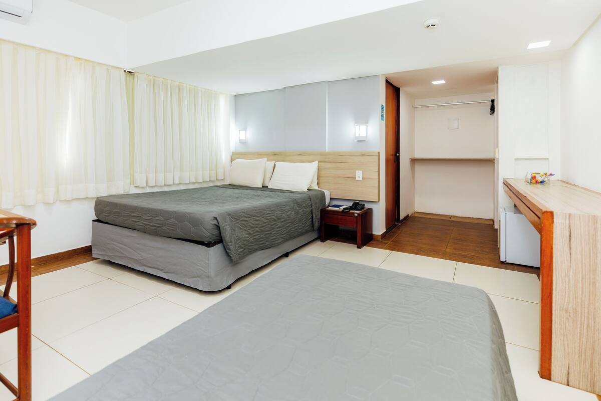 Quarto de hotel com duas camas, uma escrivaninha, uma cadeira e um pequeno espaço de cozinha.