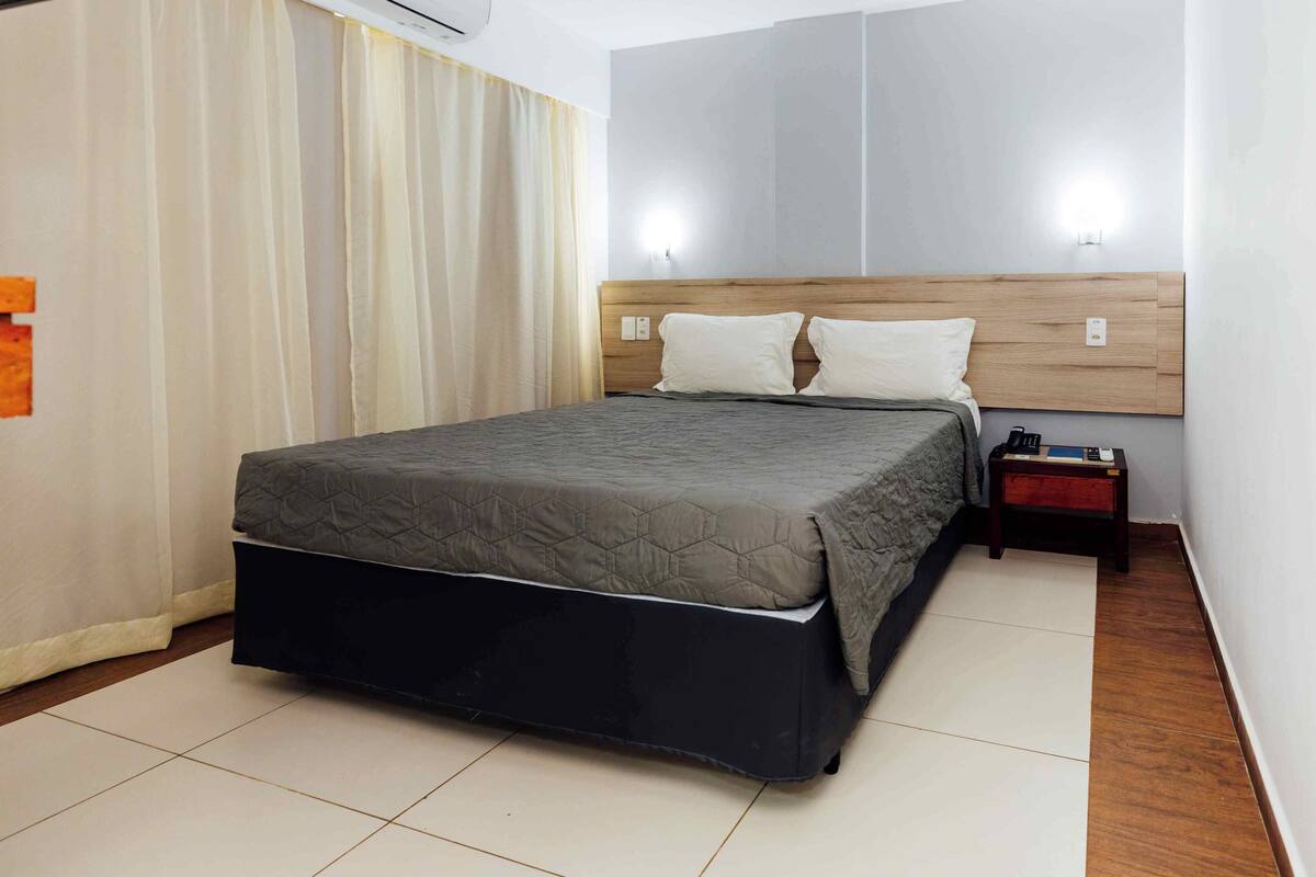 Quarto de hotel com cama, mesa de cabeceira e luminária fixada na parede.