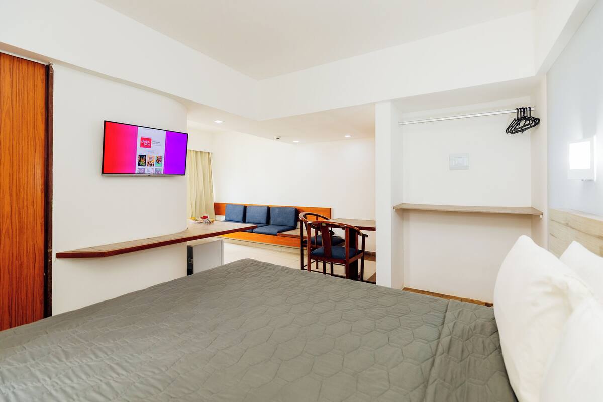 Quarto de hotel com cama, escrivaninha, cadeira e televisão fixada na parede.