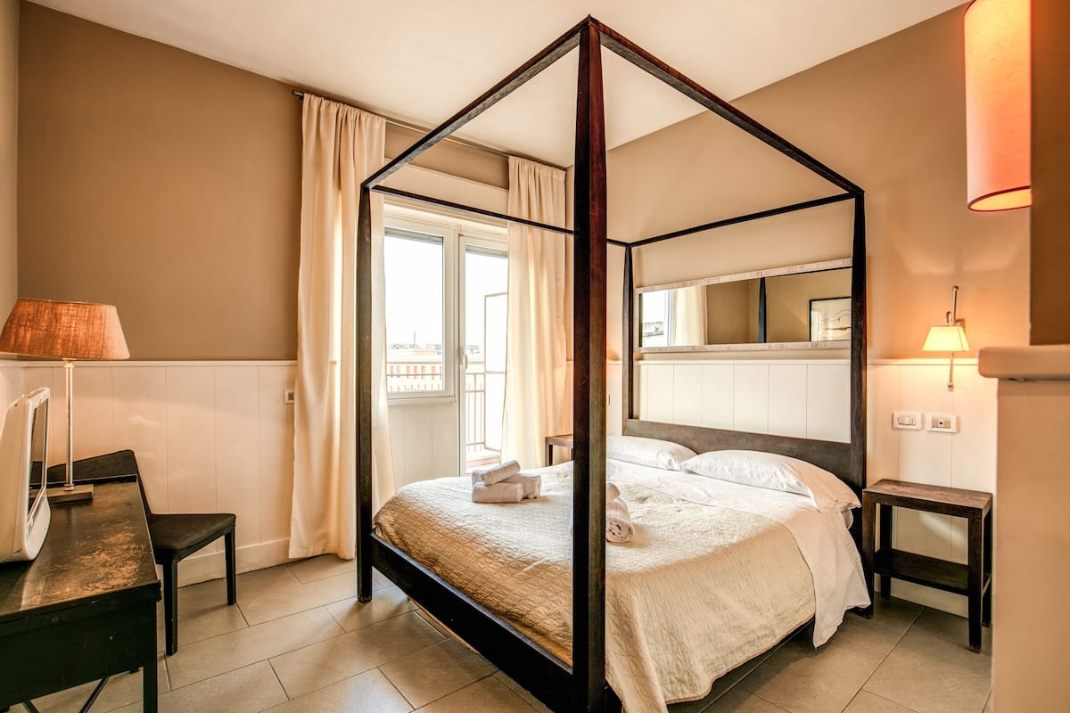 A four-poster bed with a view of the city through the window, a desk with a lamp, and a bedside table with a lamp.