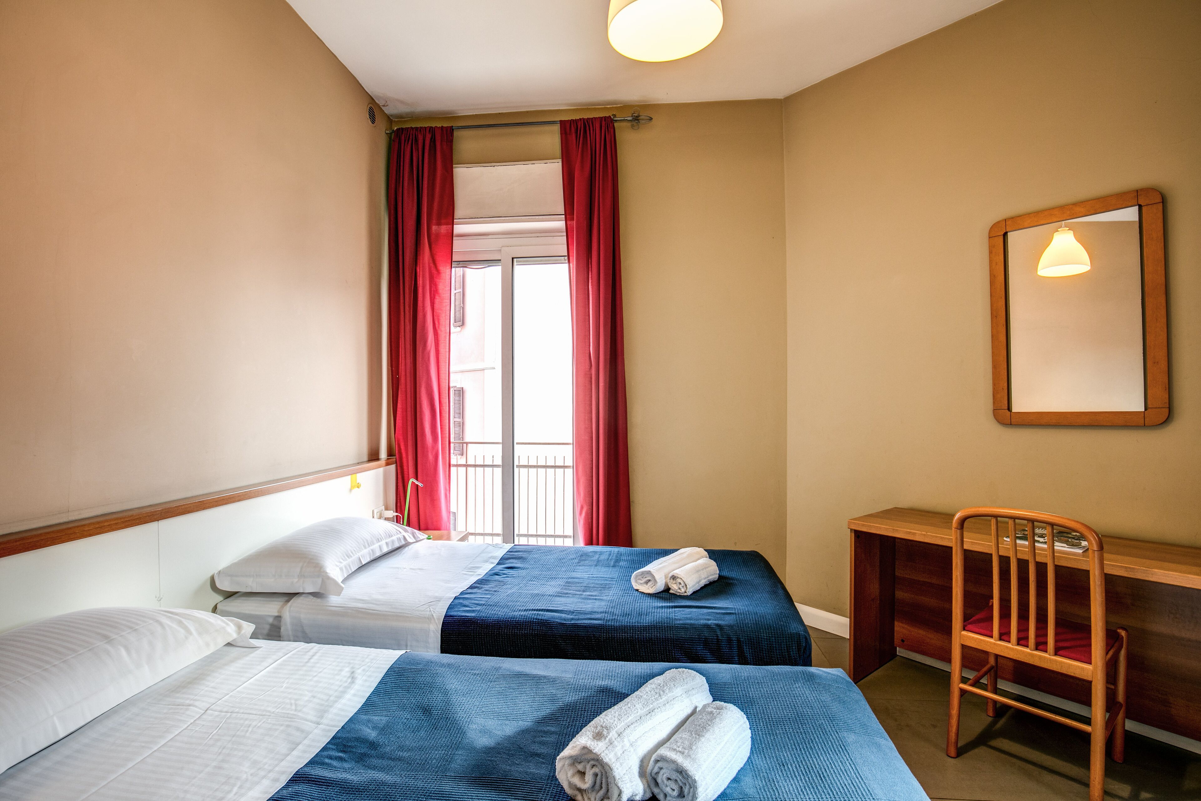 Habitación de hotel con dos camas, un escritorio, una silla y un ventanal con cortinas rojas.