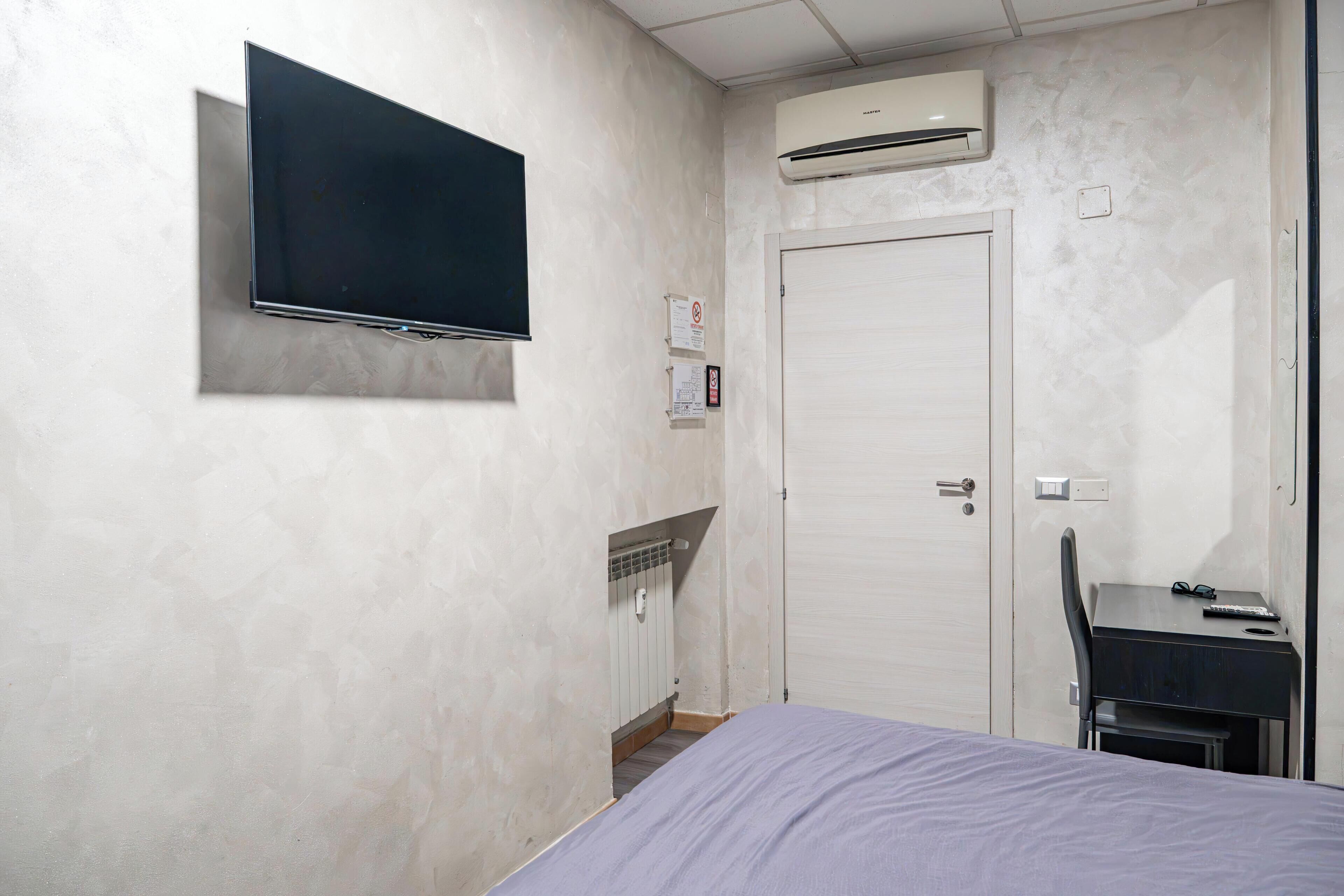 Una habitación con cama, televisor montado, aire acondicionado, puerta y escritorio con silla.