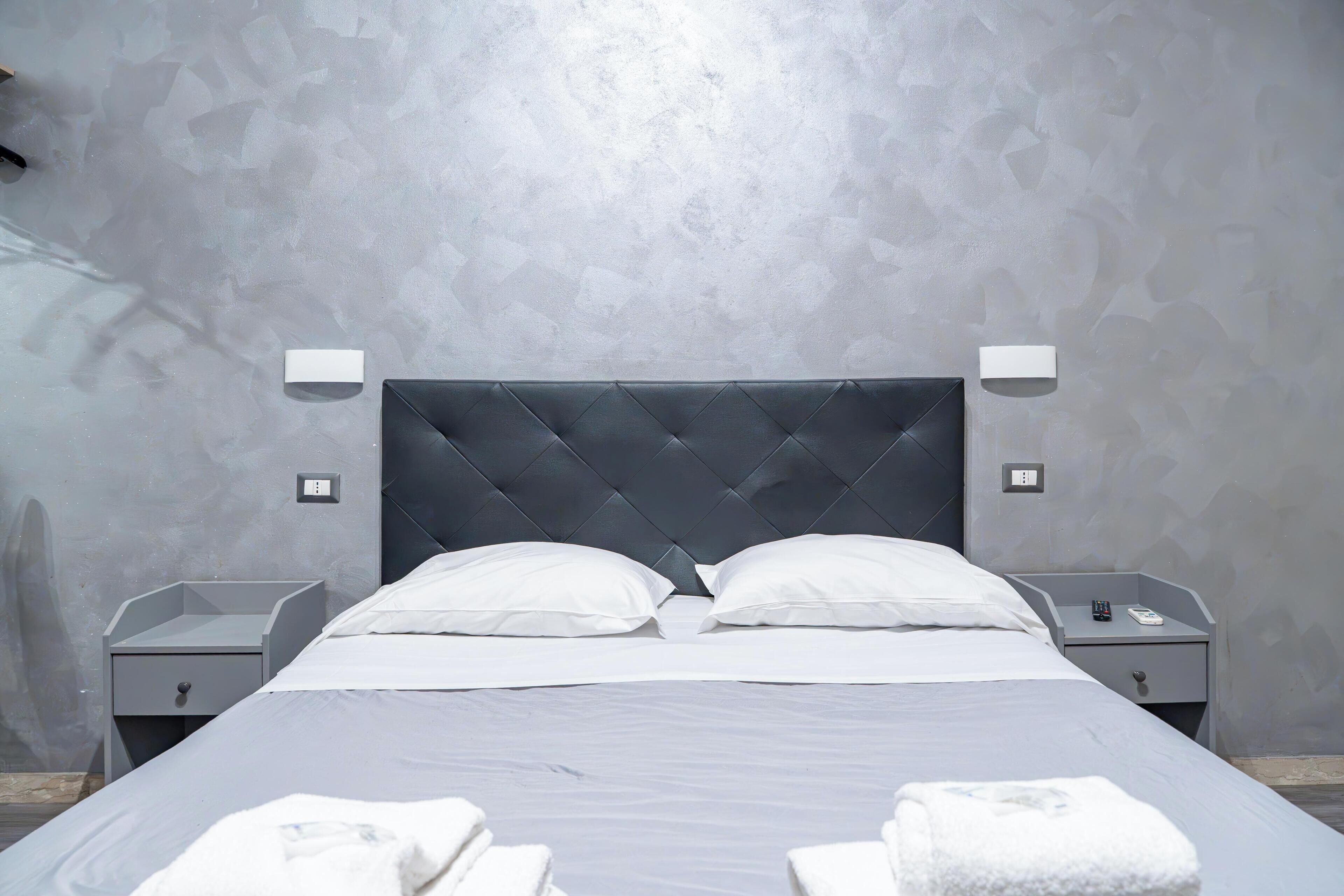 Una habitación de hotel moderna con una cama grande, dos mesitas de noche y una pared gris con textura.