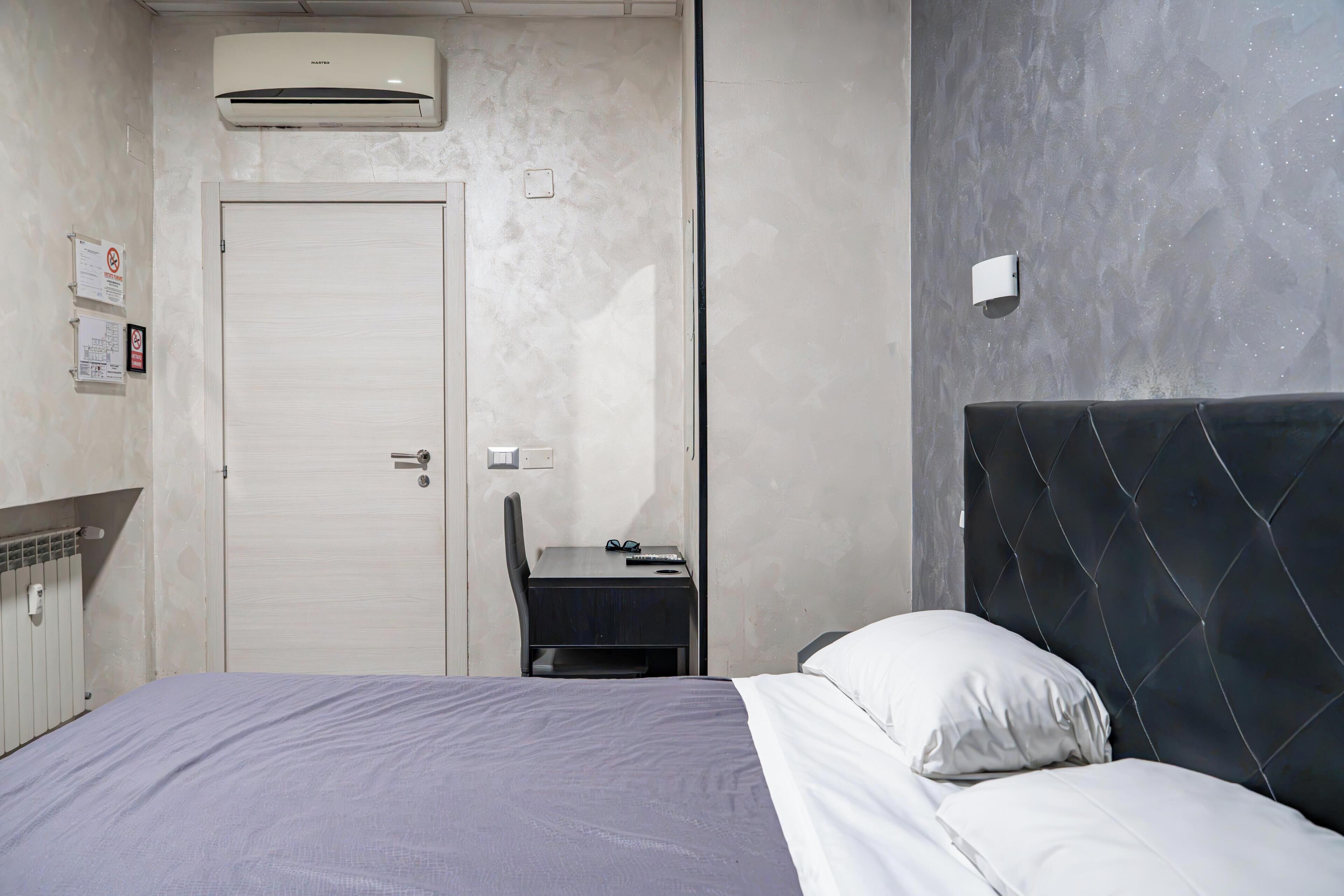 Un dormitorio con cama, escritorio y silla. Se observa una puerta, un equipo de aire acondicionado y una lámpara fijada a la pared.