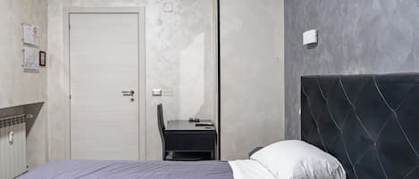 Un dormitorio con cama, escritorio y silla. Se observa una puerta, un equipo de aire acondicionado y una lámpara fijada a la pared.