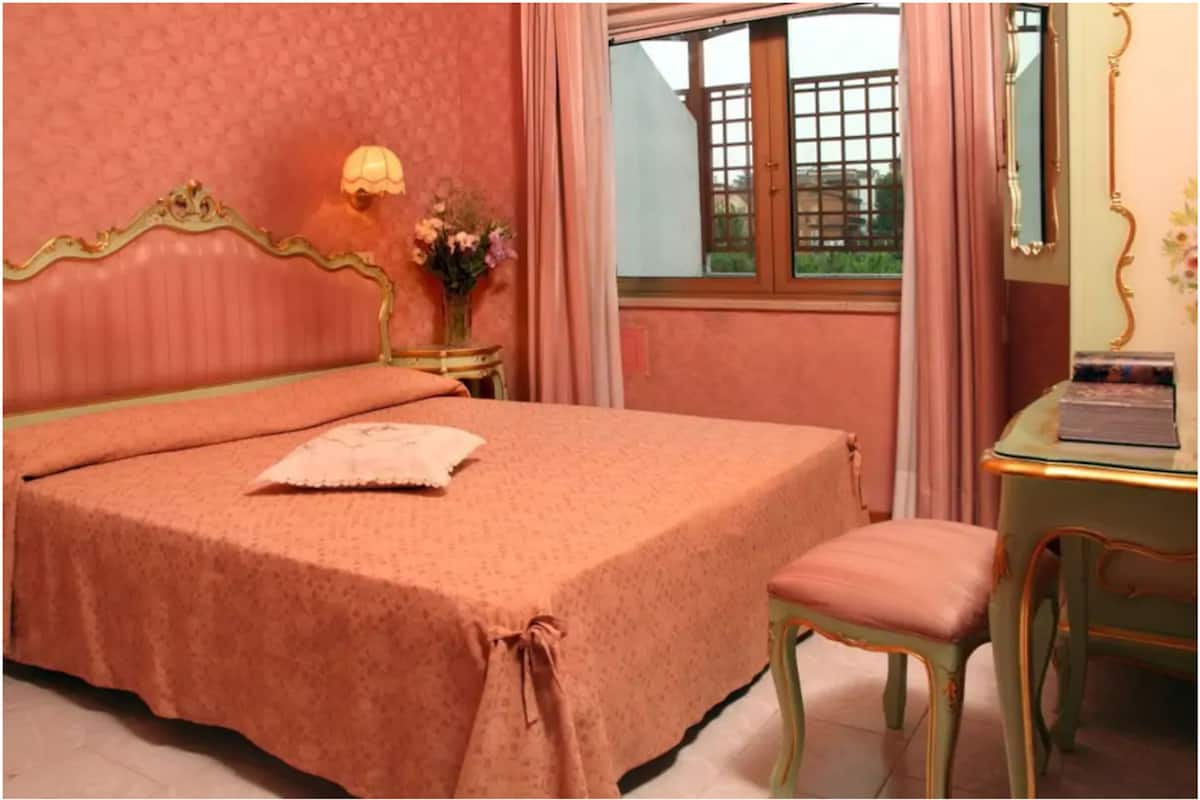 Una habitación con una cama, una mesita con una lámpara, una silla y un ventanal con cortinas.