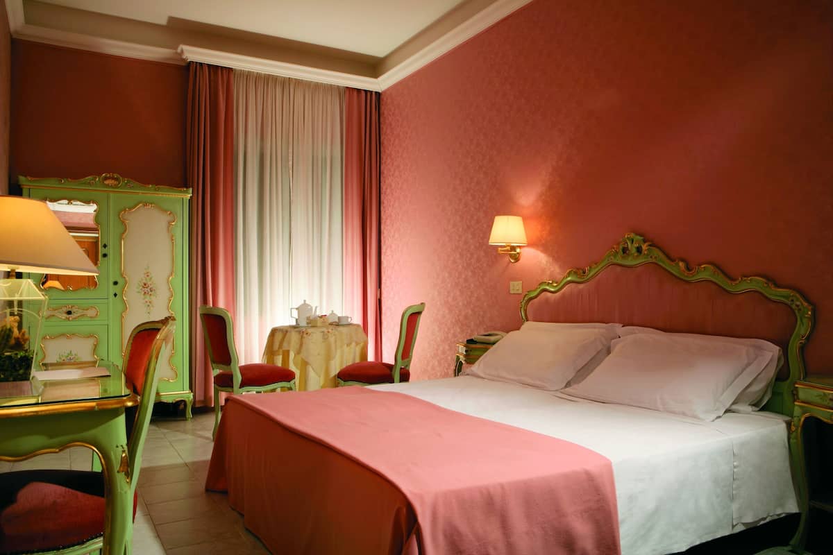 Habitación de hotel con cama, escritorio con lámpara, una silla, una mesita con lámpara y una ventana con cortinas.