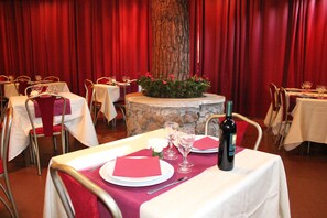 A restaurant setting with tables set for dining, featuring red napkins, wine glasses, and a bottle of wine.