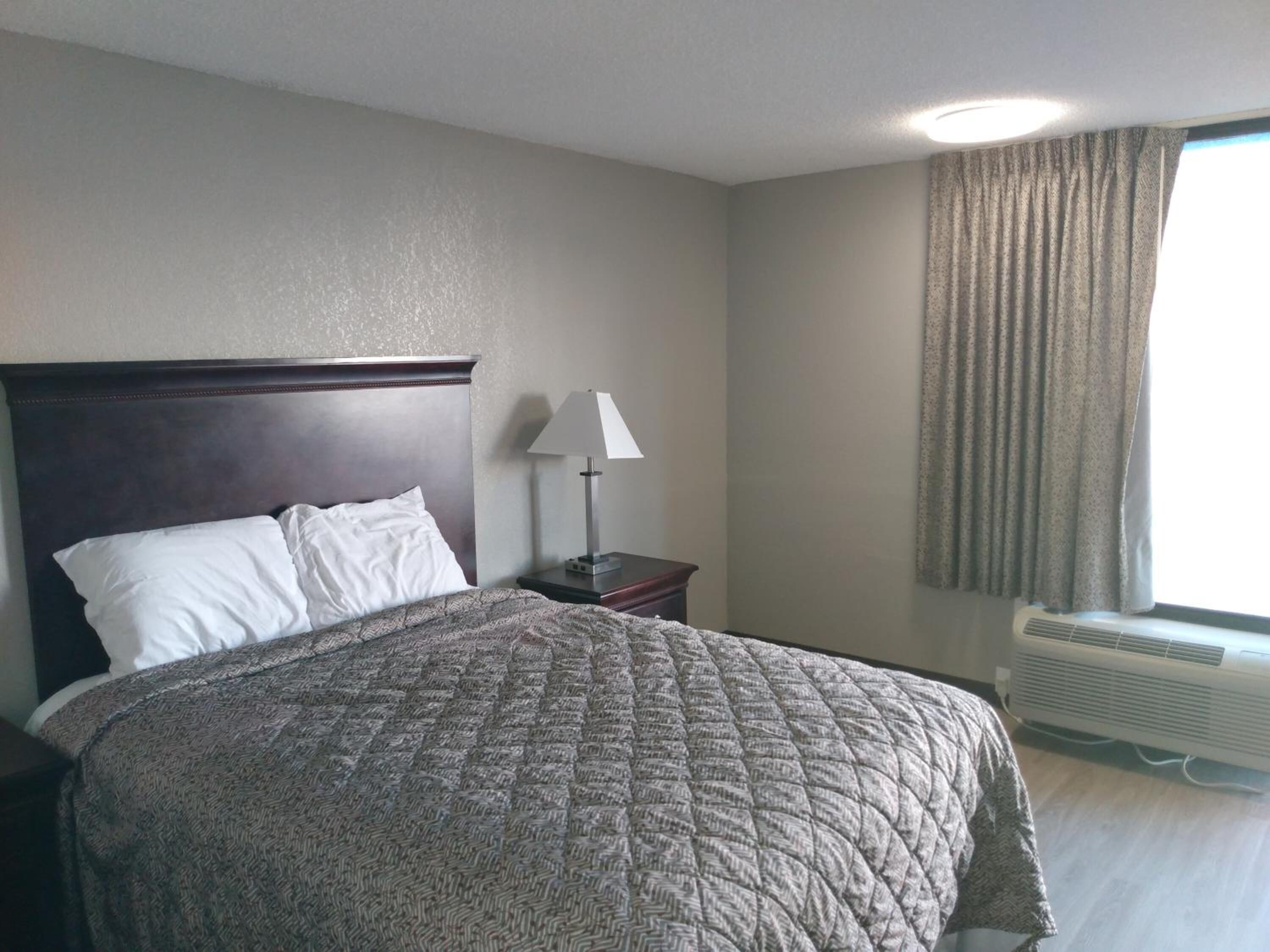 A hotel room with a large bed, a nightstand with a lamp, a window with curtains, and an air conditioning unit.
