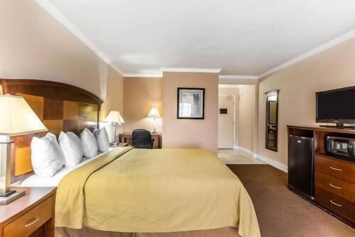 A hotel room with a bed, bedside lamps, a desk with a lamp, a television, a dresser, and a framed picture on the wall.