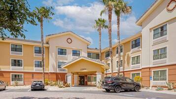 A hotel with a yellow facade, a 'Lobby' sign, palm trees, and a parking lot with cars.